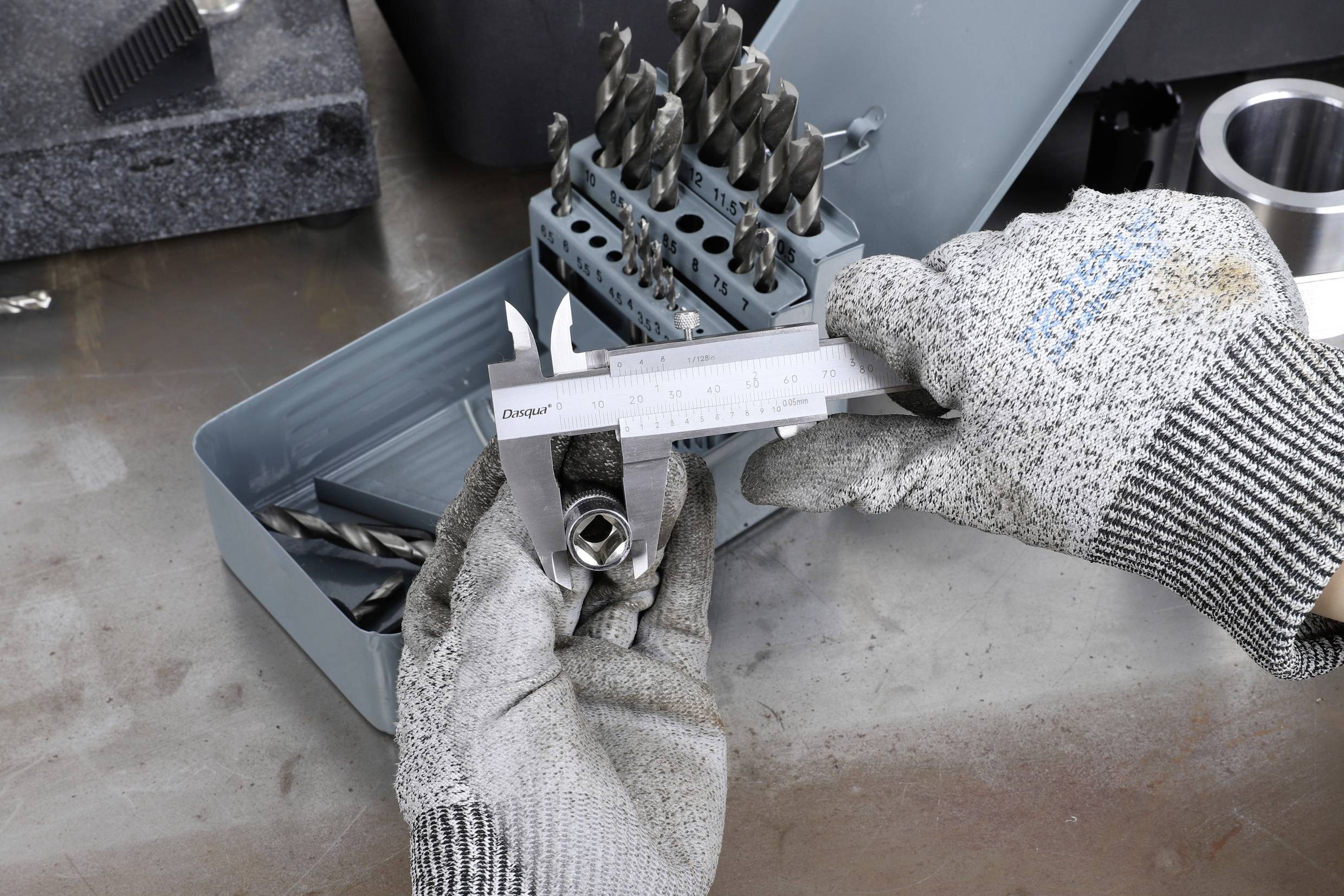 Une personne portant des gants gris tient un pied à coulisse et mesure un objet métallique, tandis que des forets en métal sont rangés dans une boîte à côté.