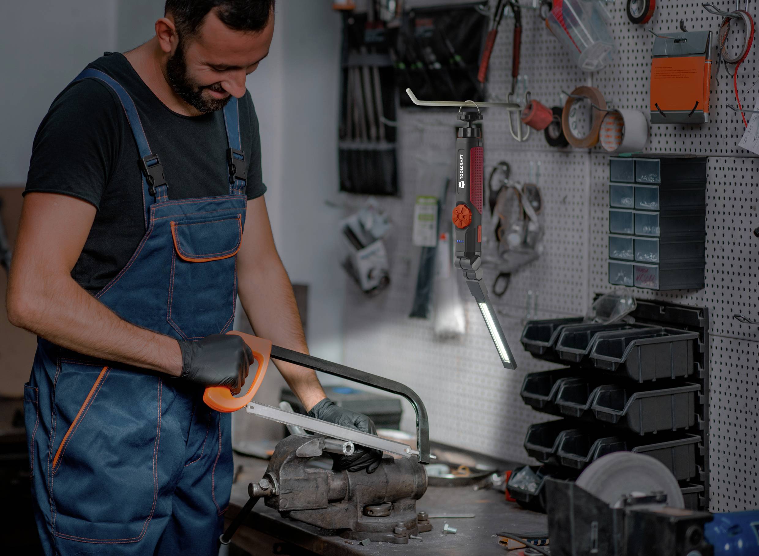 Un homme en vêtements de travail scie un tuyau métallique avec une scie à main dans un atelier. Des outils sont accrochés au mur.