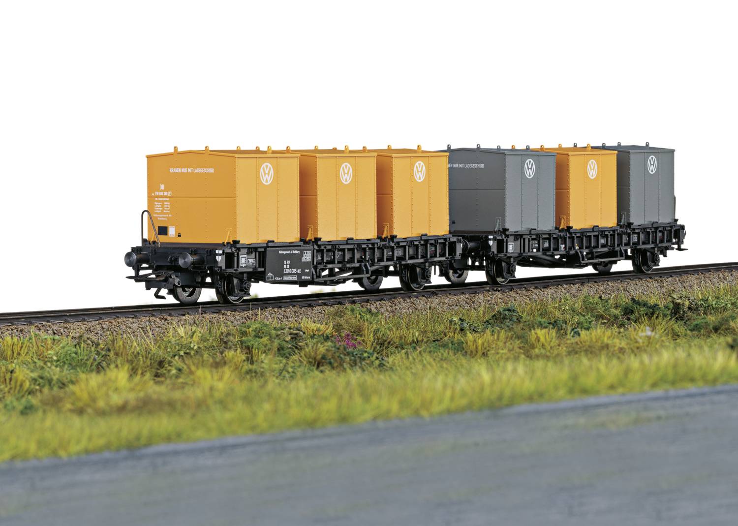 Un train de marchandises de chemin de fer miniature avec des conteneurs orange et gris circule sur un rail à travers un paysage verdoyant.