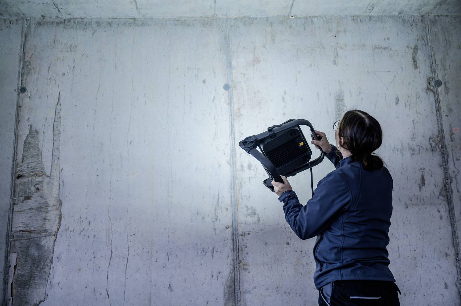 Une personne éclaire un mur de béton gris avec un projecteur portable dans une pièce apparemment inachevée.