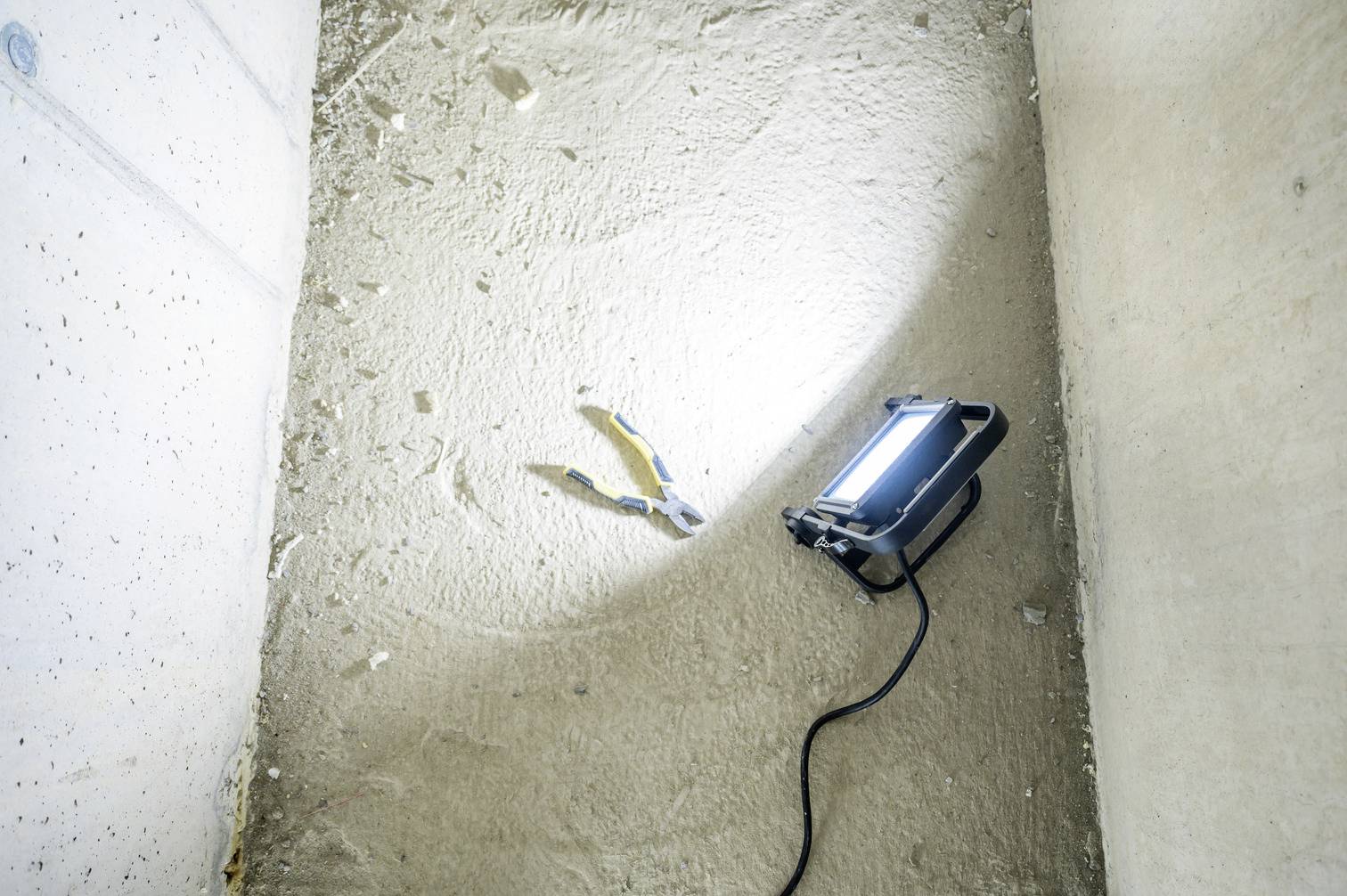 Un projecteur de chantier éclaire des pinces sur un sol désolé et poussiéreux dans une pièce claire et fermée.
