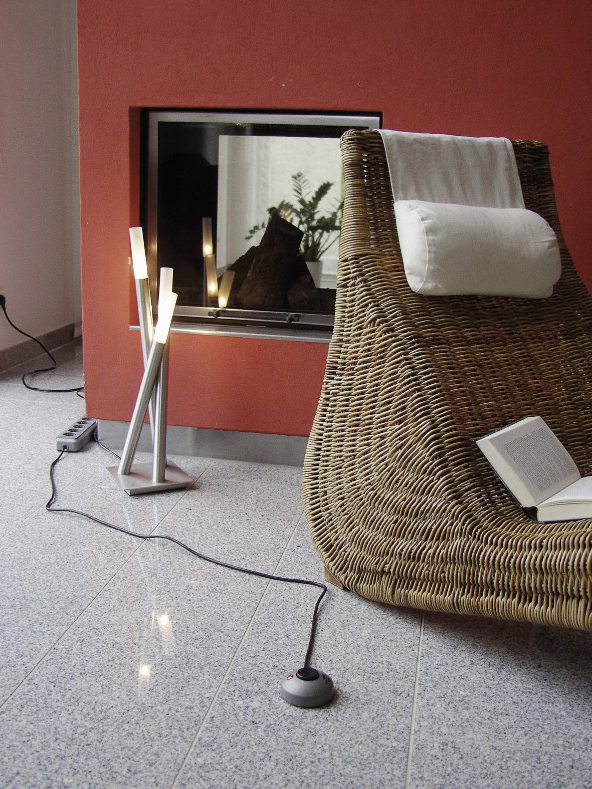 Un fauteuil tressé est placé devant une cheminée sur un mur rouge. Un livre ouvert est posé sur le sol, à côté d'une lampe moderne.