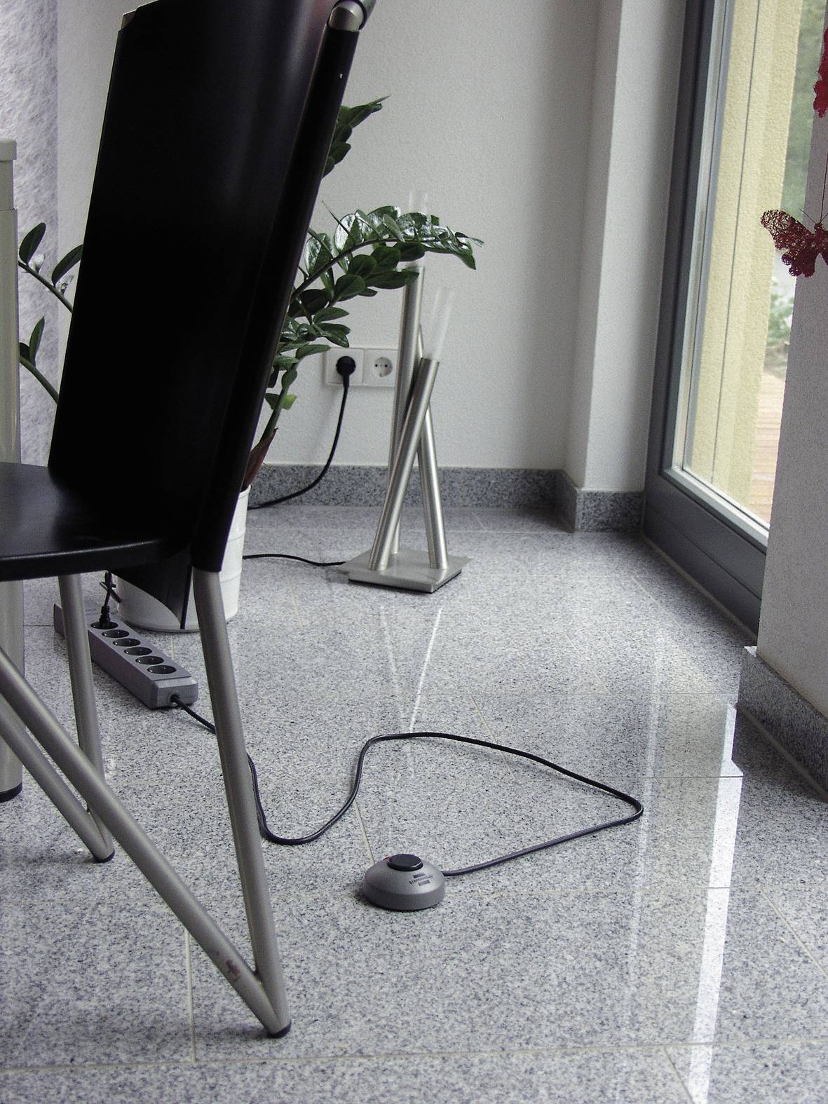 Une chaise et une plante sont placées sur un sol en pierre brillant, à côté d'une porte vitrée. Un câble avec un interrupteur au pied est posé sur le sol.