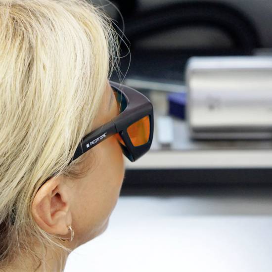 Une personne aux cheveux blonds porte des lunettes de protection spéciales avec des verres oranges. Des appareils sont flous en arrière-plan.