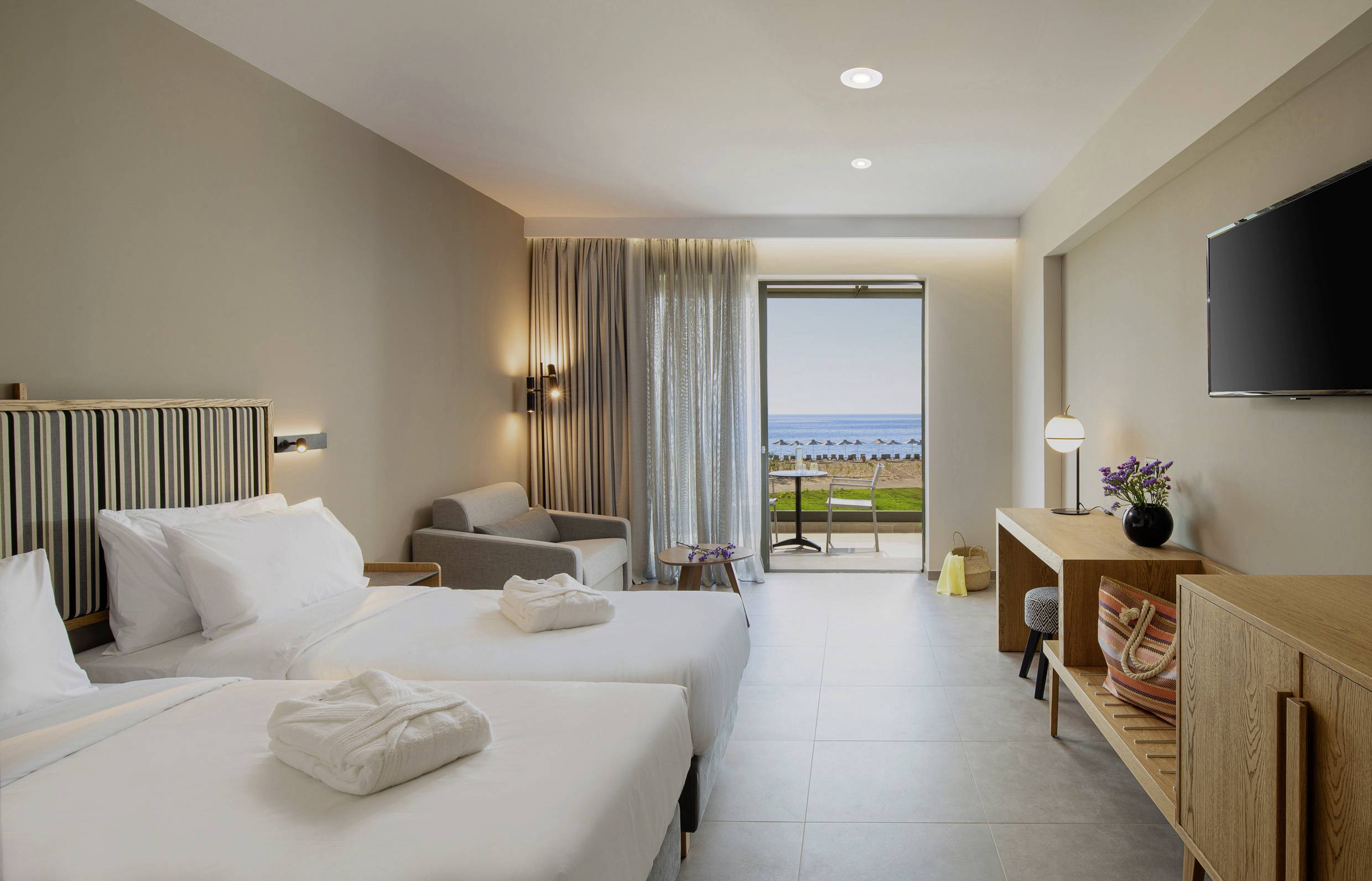 Chambre d'hôtel avec deux lits simples, un canapé, une table, une télévision et un balcon offrant une vue sur la mer. Décoration lumineuse et moderne.