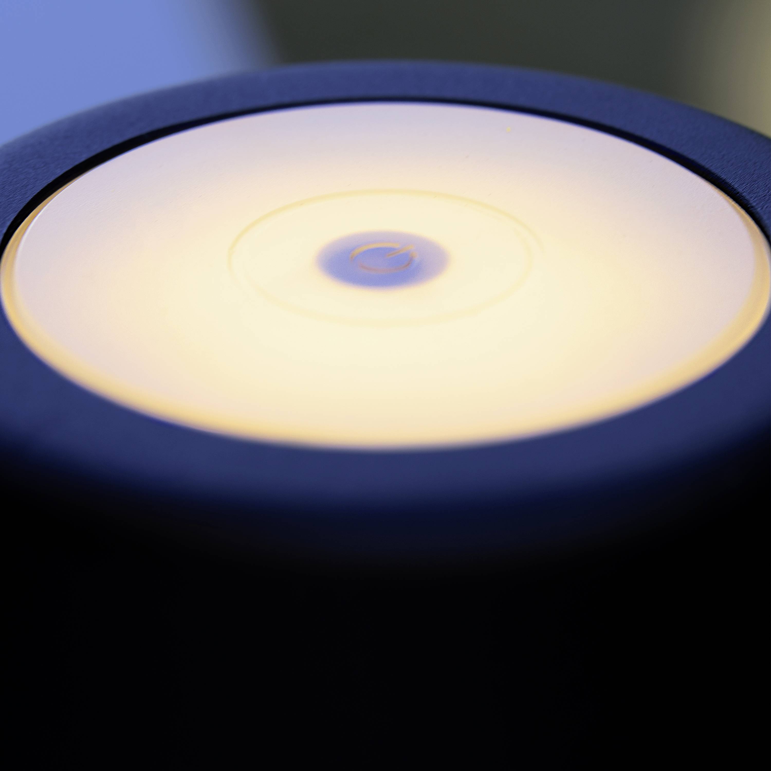 Une lampe de table avec un abat-jour circulaire lumineux, probablement à LED, dans une pièce tamisée, crée une atmosphère chaleureuse.