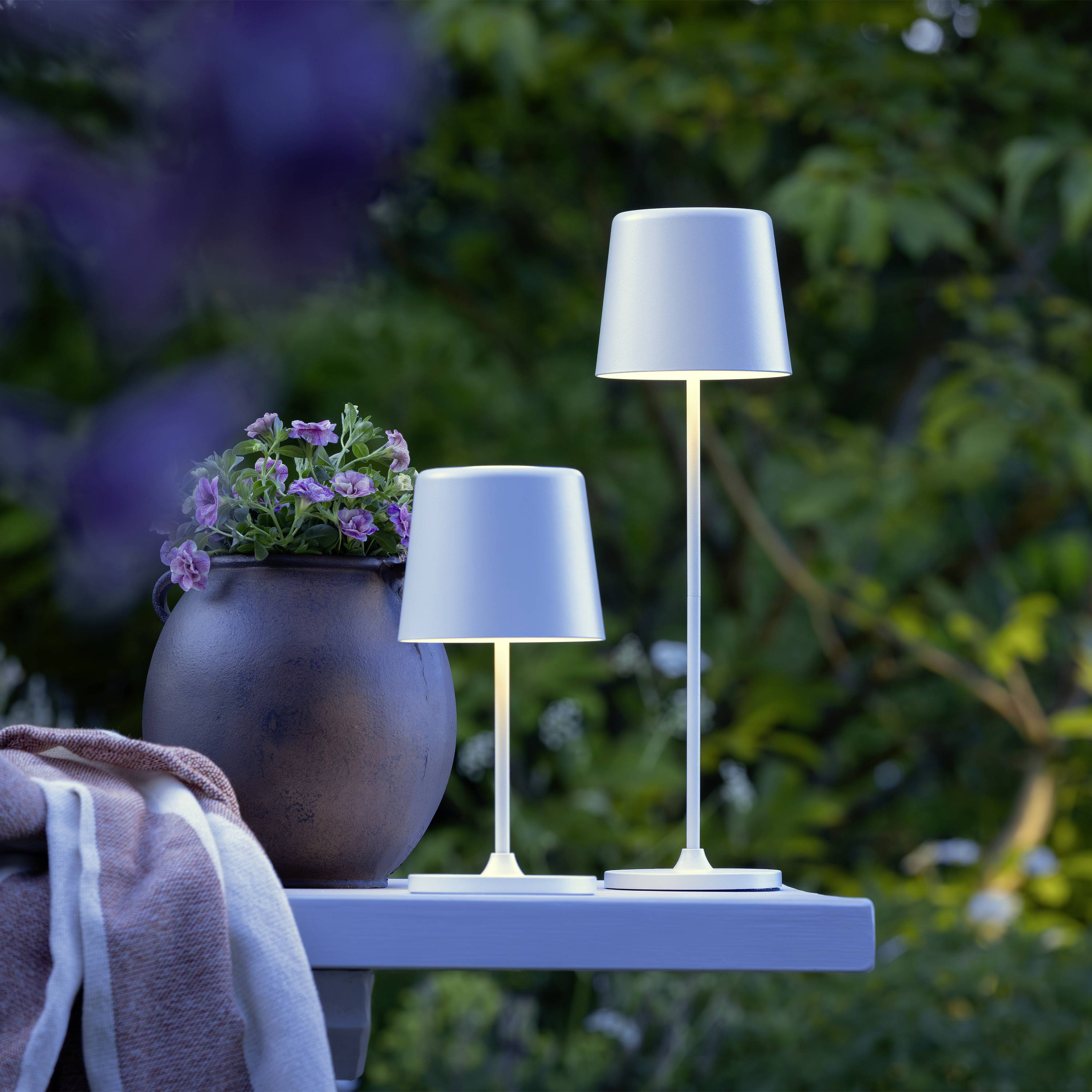 Deux lampes de table modernes et un pot de fleurs avec des fleurs violettes sont placés sur une table de jardin à l'extérieur, avec un arrière-plan vert flou.