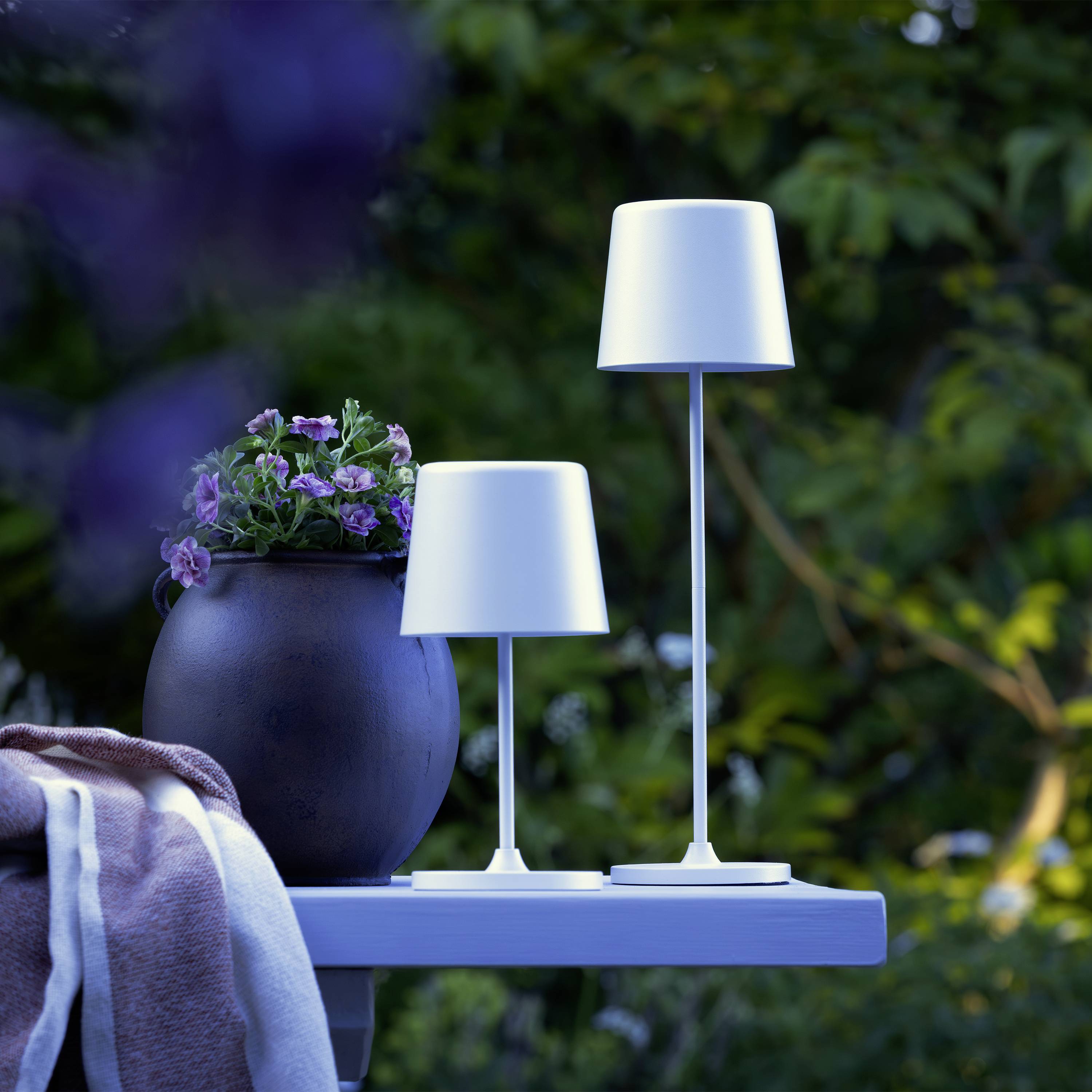 Une table de jardin dressée à l'extérieur avec deux lampes de table élégantes et modernes, et un pot contenant des fleurs mauves, entourée de plantes.