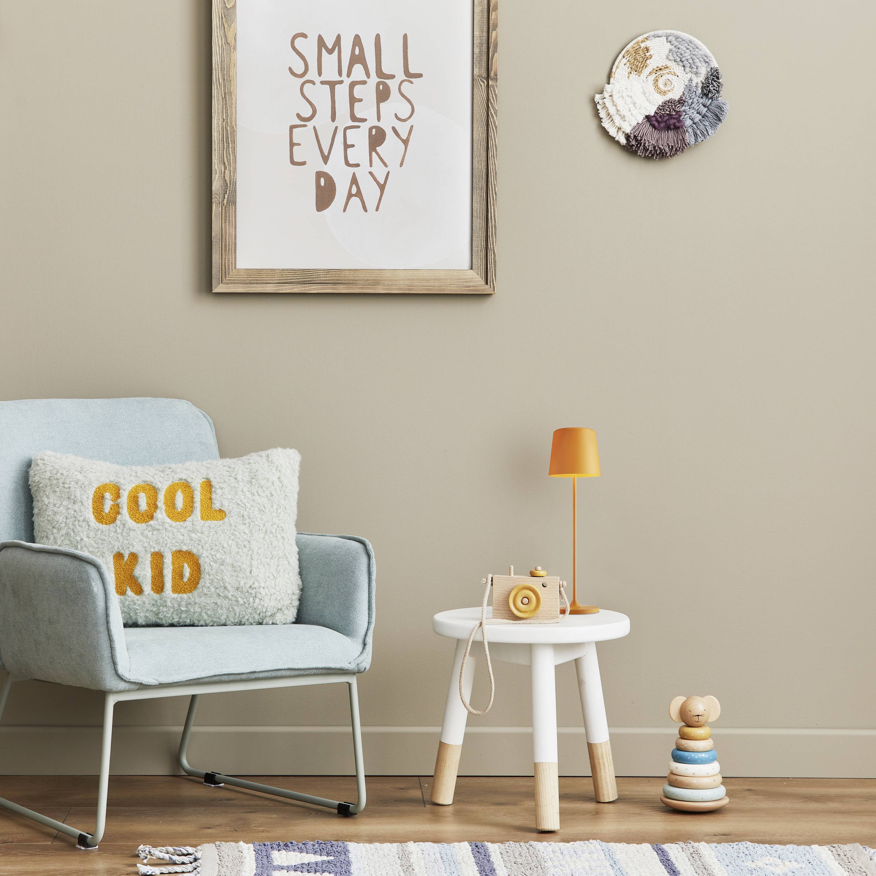 Une chambre d'enfant moderne avec un fauteuil bleu et un coussin "COOL KID", une table d'appoint blanche avec une lampe jaune, une décoration murale et un tableau encadré portant l'inscription "SMALL STEPS EVERY DAY".