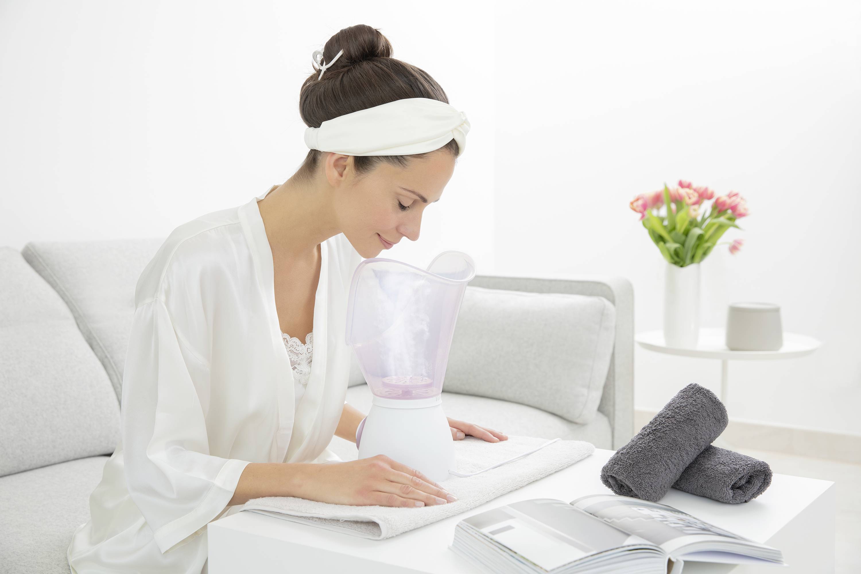 Une femme est assise sur le canapé et utilise un vaporisateur facial. À côté d'elle se trouvent des serviettes et un livre ouvert. À l'arrière-plan, il y a un vase avec des fleurs.