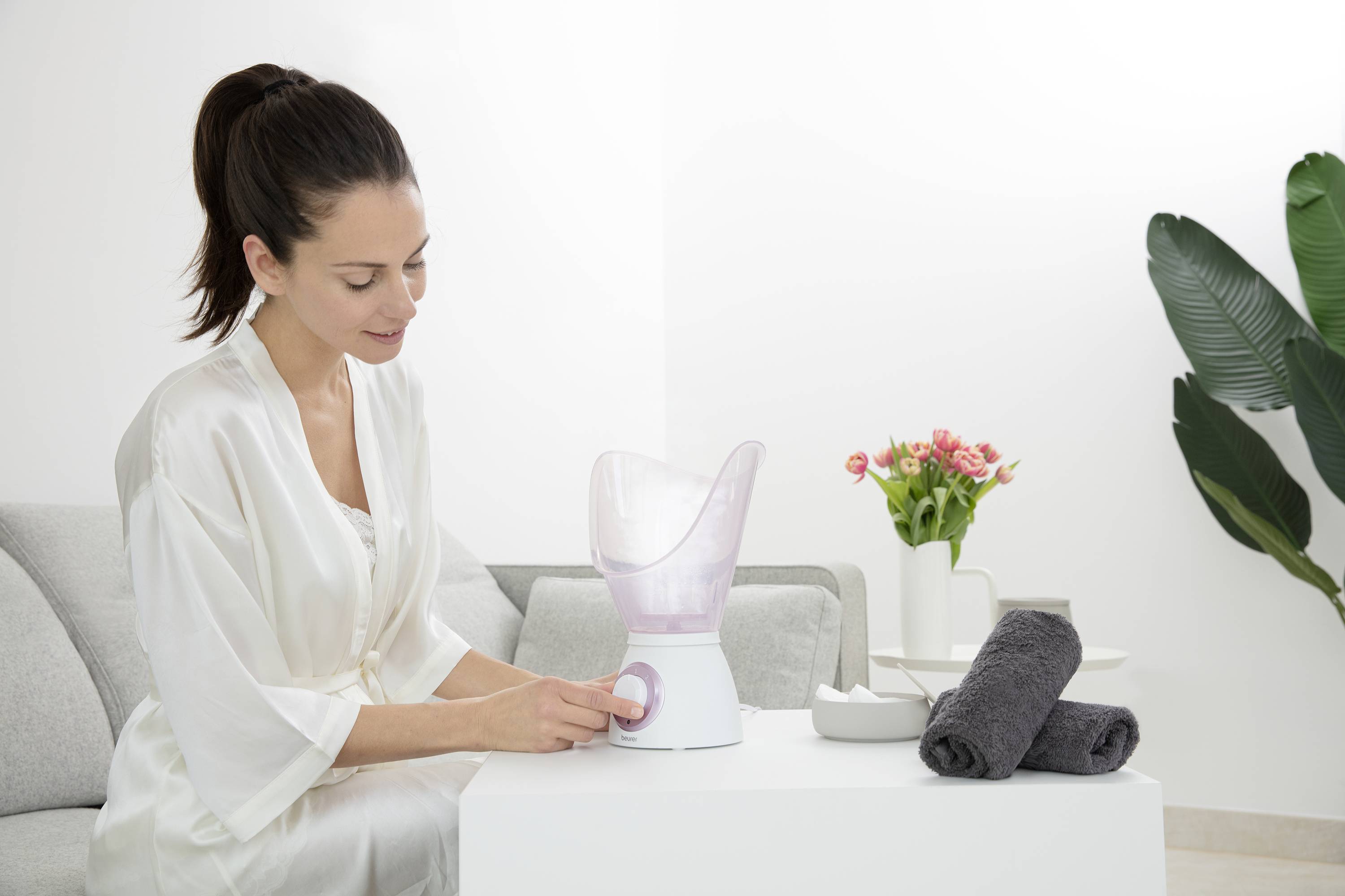 Une femme est assise sur le canapé et utilise un vaporisateur facial. À côté d'elle se trouvent des serviettes et un vase rempli de fleurs.