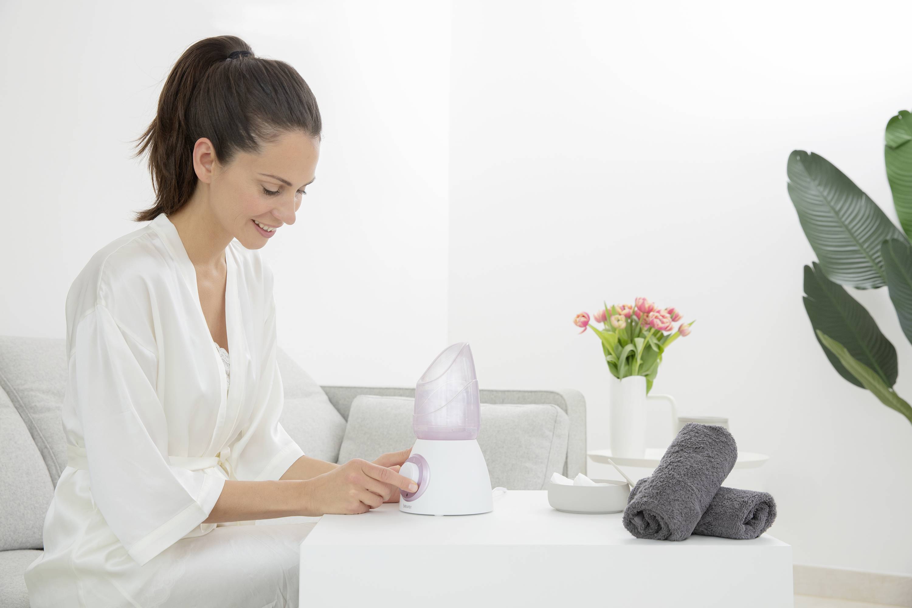 Une femme utilise un appareil de vapeur faciale dans un salon lumineux. À côté d'elle, des serviettes et des fleurs sont disposées sur une table.