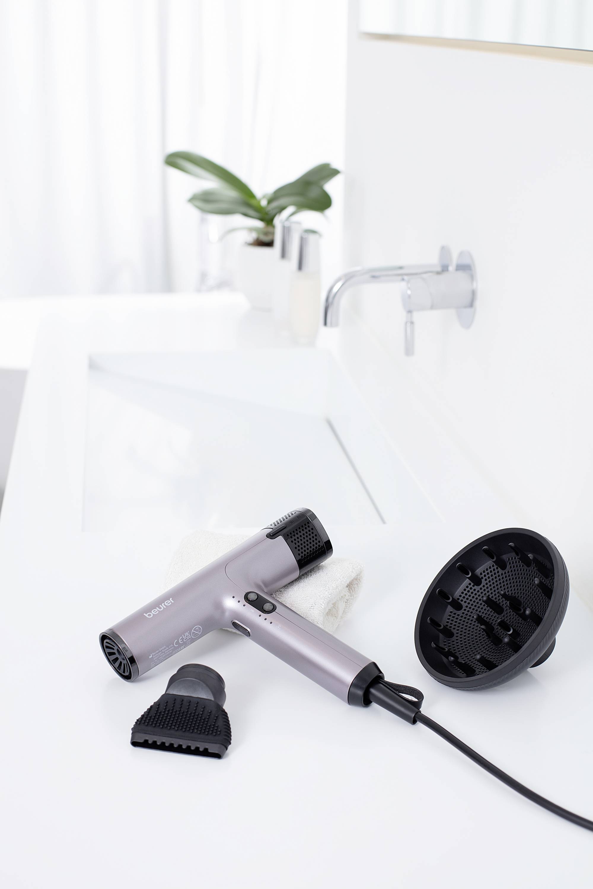 Un sèche-cheveux gris avec différents accessoires est posé sur le lavabo blanc. Des plantes sont visibles en arrière-plan.