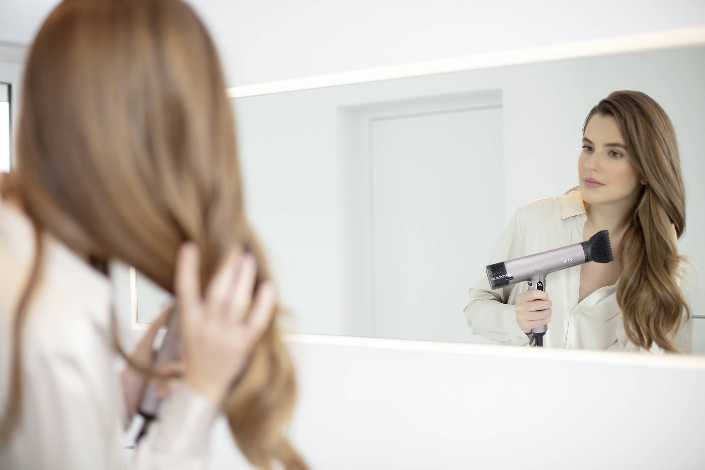 Une femme sèche ses longs cheveux bruns devant un miroir dans une pièce lumineuse. Elle porte un haut blanc.