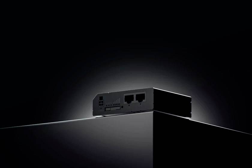 Un petit appareil réseau noir avec deux ports Ethernet et des voyants lumineux est posé sur un socle sombre devant un fond noir.