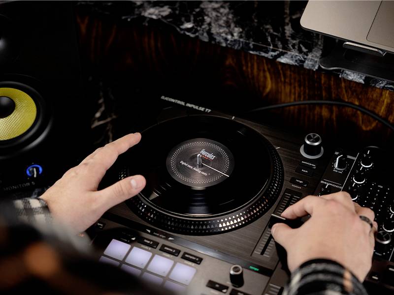 Une personne pose un disque vinyle sur un contrôleur DJ. Des enceintes sont visibles en arrière-plan.