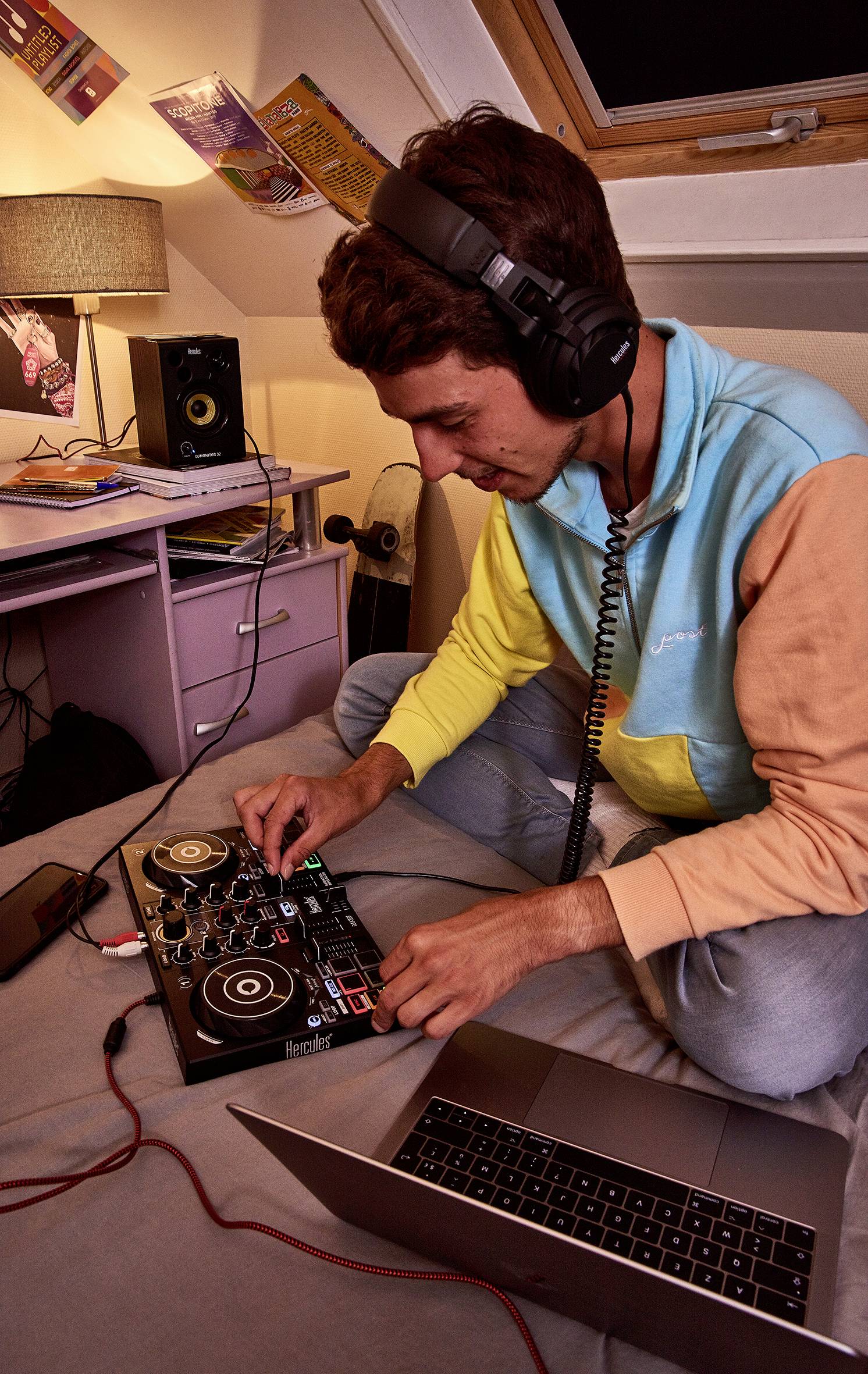 Une personne est assise sur un lit et mixe de la musique avec un contrôleur DJ. Elle porte des écouteurs et un sweat de couleur. Un ordinateur portable est posé en arrière-plan.