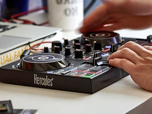 Une personne utilise une table de mixage DJ Hercules avec plusieurs boutons et curseurs. Un ordinateur portable et une tasse sont visibles en arrière-plan sur une table.