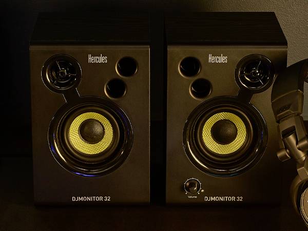 Deux enceintes noires avec des membranes jaunes et l'inscription 'Hercules DJMonitor 32' sur la face avant. Un casque audio est posé à côté à droite.