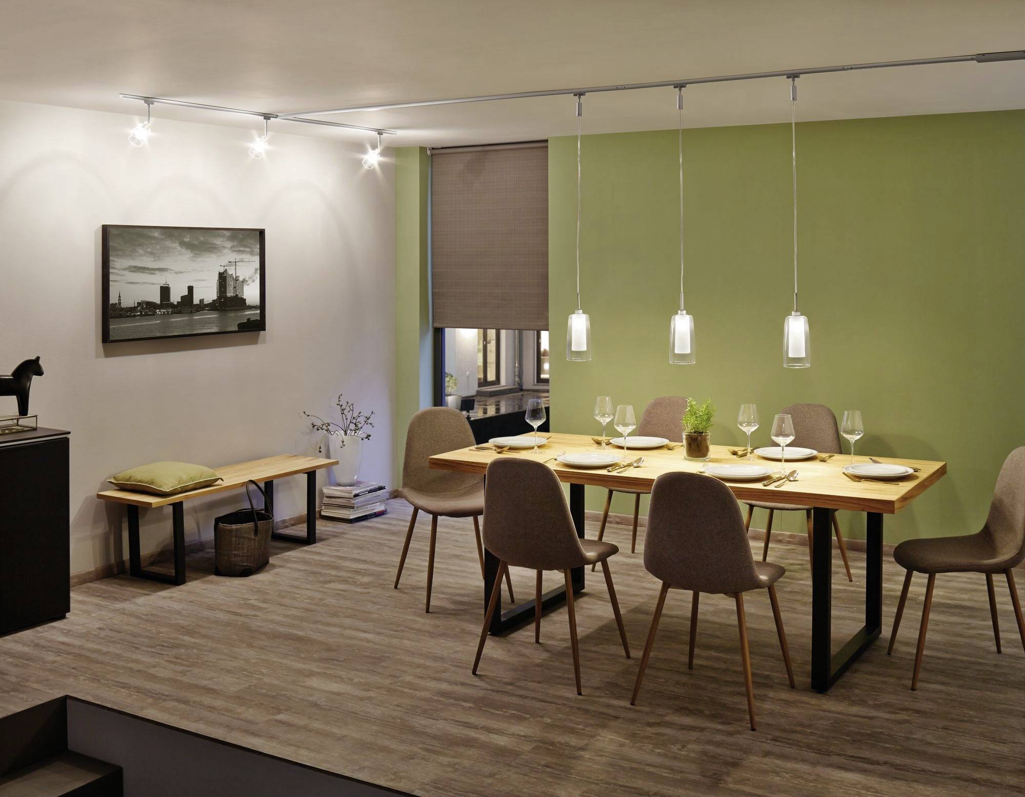 Salle à manger avec une table en bois, six chaises et un système d'éclairage moderne. Un mur vert et une œuvre d'art créent une atmosphère chaleureuse.