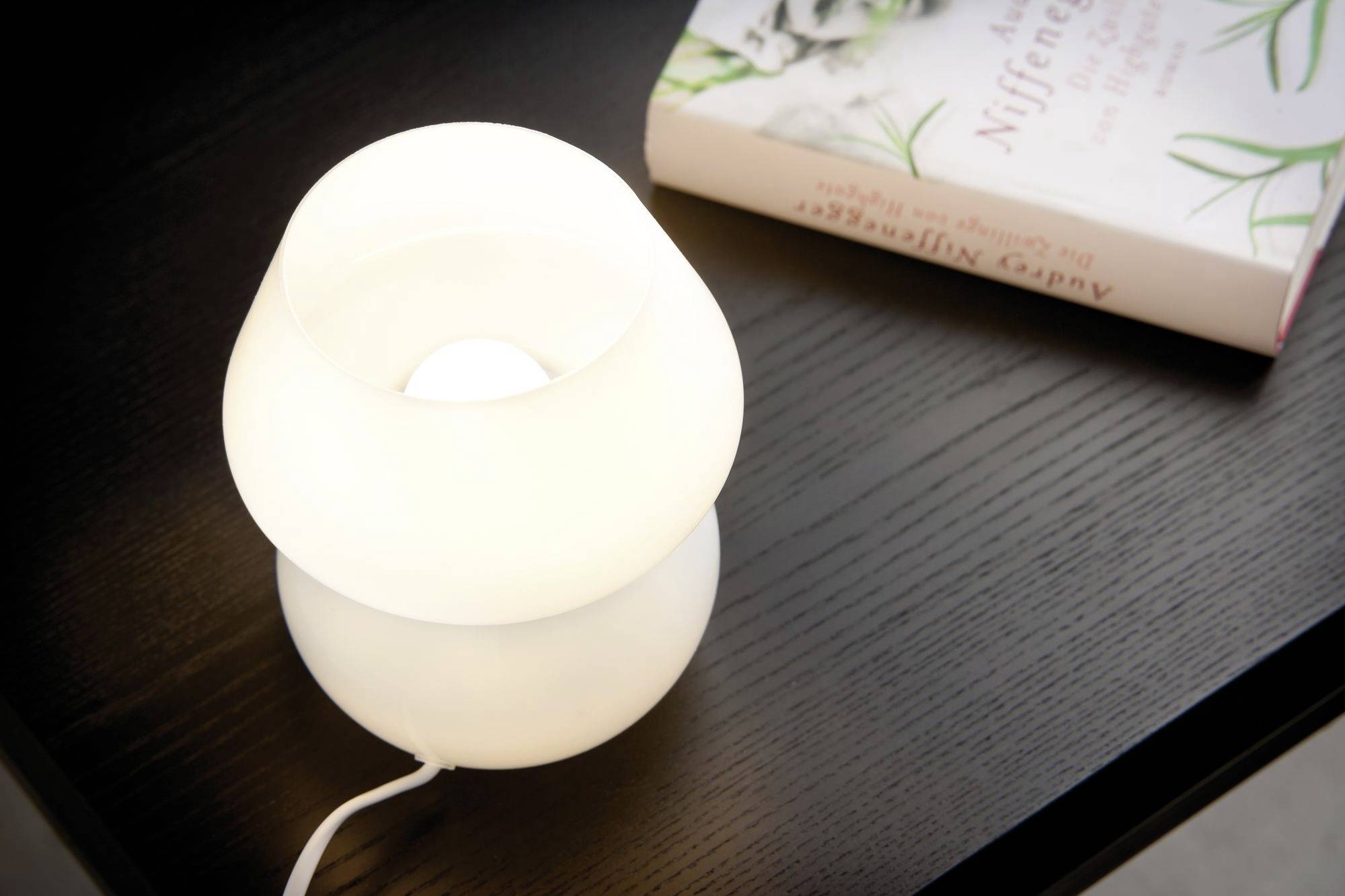 Une lampe de table blanche s'allume sur une table en bois sombre, à côté d'un livre dont la couverture présente un motif botanique.