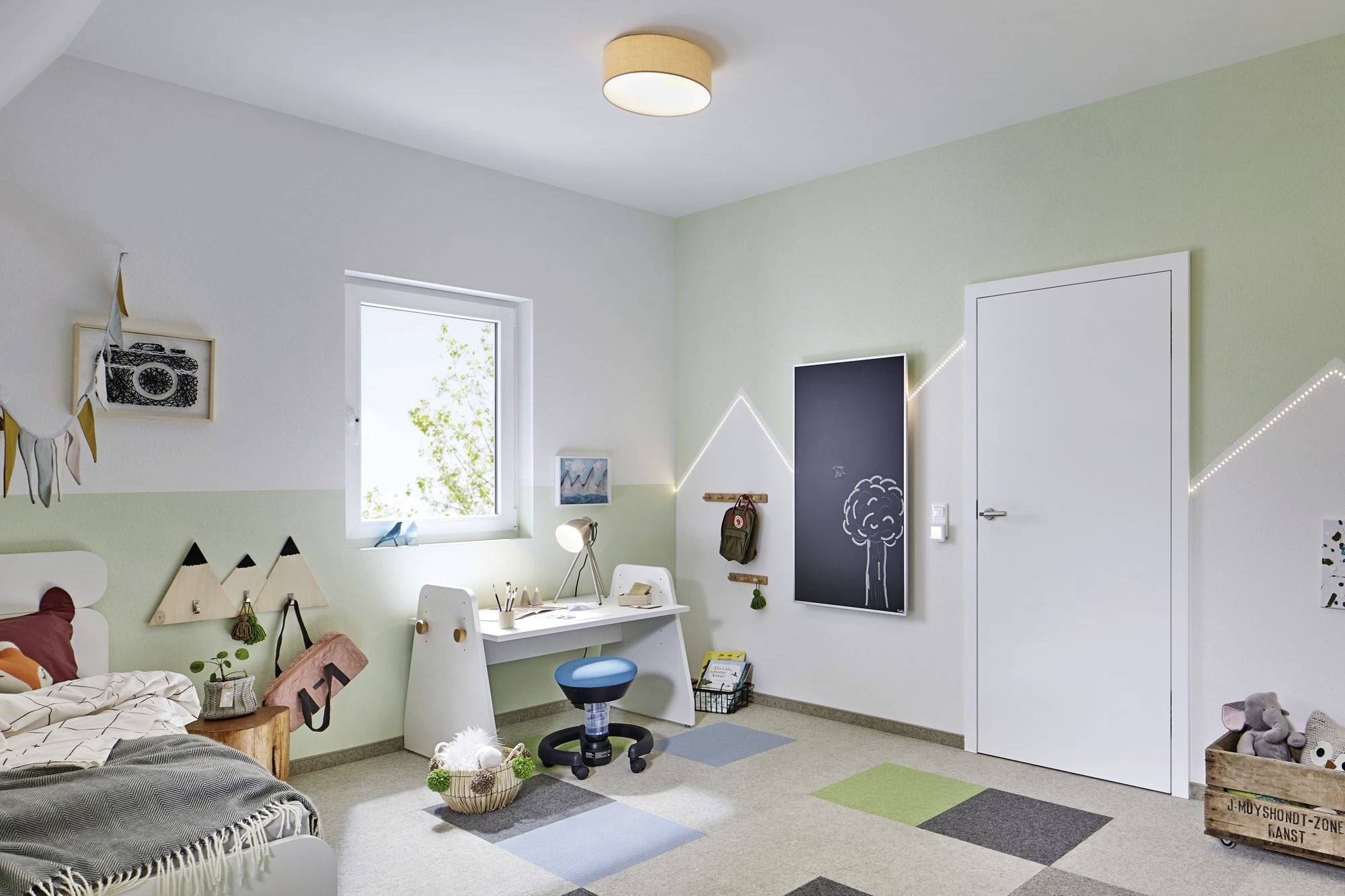 Une chambre d'enfant aux murs vert pastel. À gauche se trouve un lit, à droite un bureau avec une chaise et des jouets. Atmosphère lumineuse et accueillante.