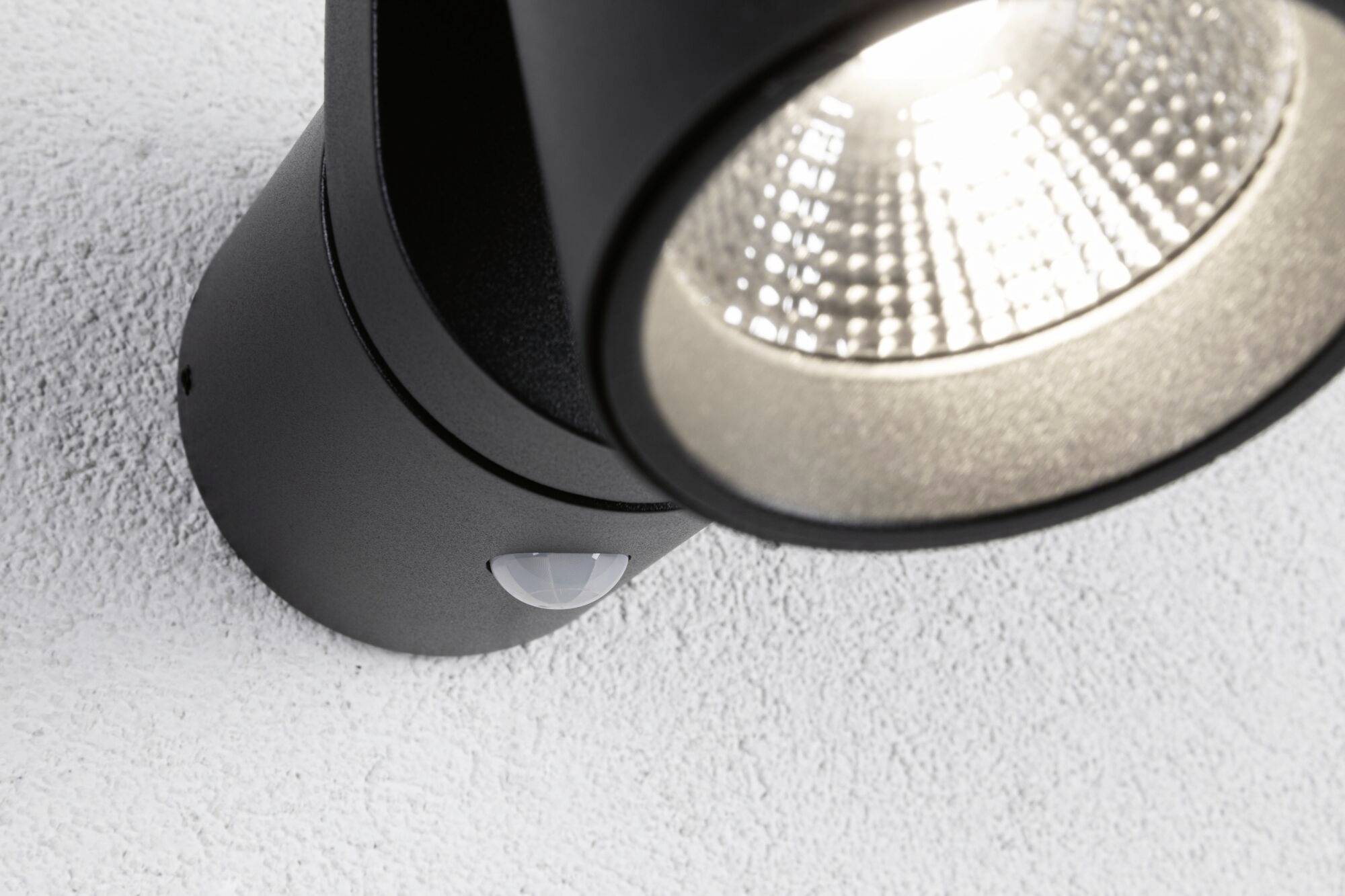 Projecteur cylindrique noir avec détecteur de mouvement, projetant une lumière vive sur une surface blanche. Idéal pour l'éclairage intérieur moderne.