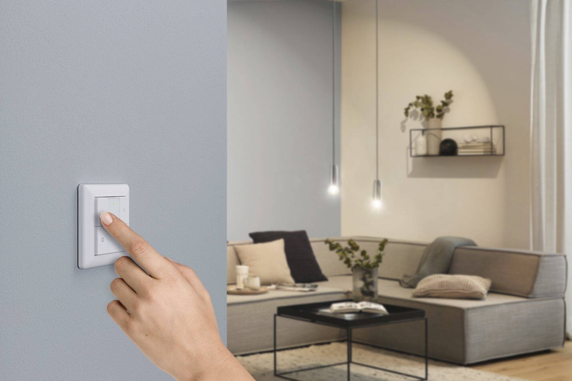 Une main allume la lumière dans un salon moderne avec un canapé gris et des lampes suspendues.
