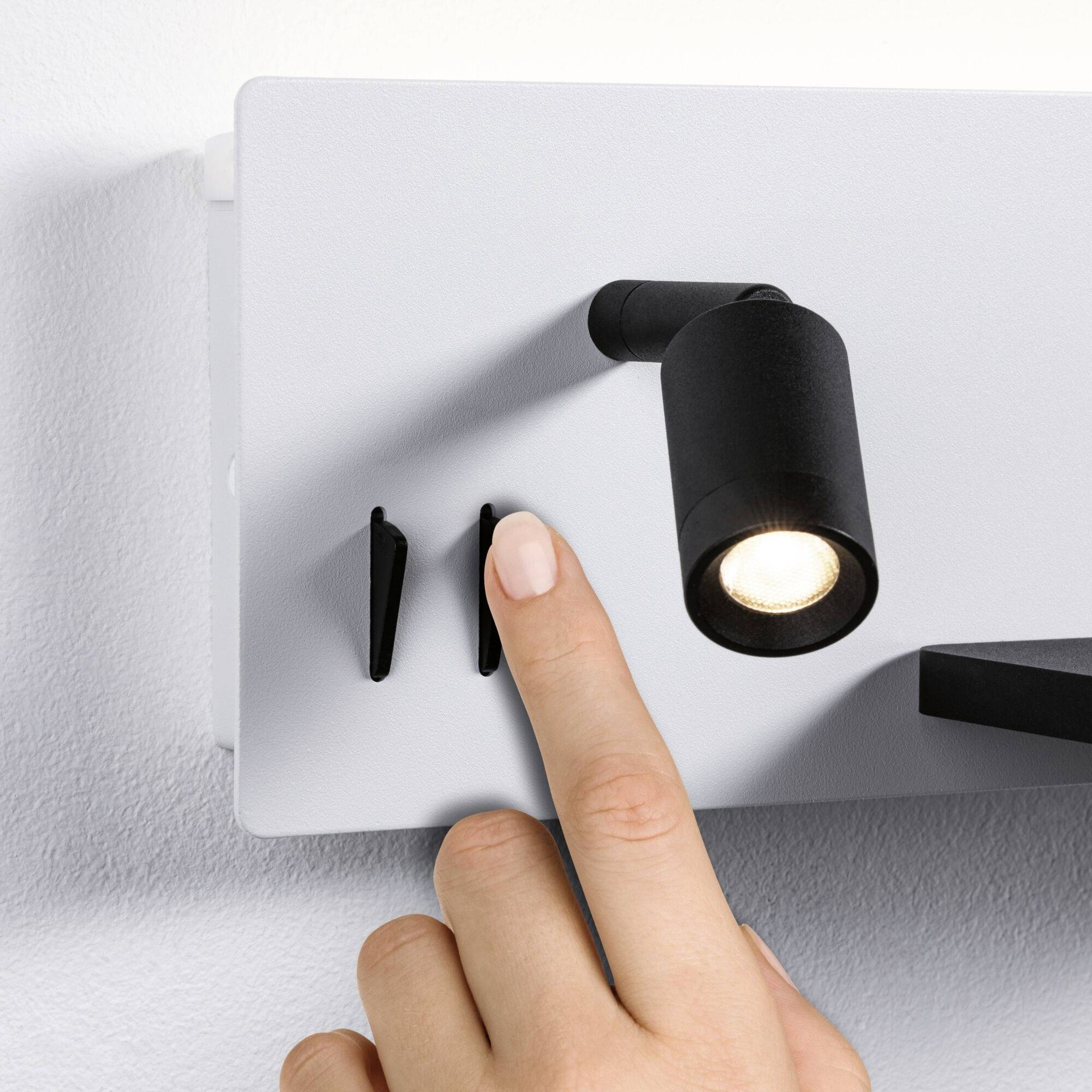 Une main allume un applique murale. La lampe est moderne et possède une tête de lampe orientable.