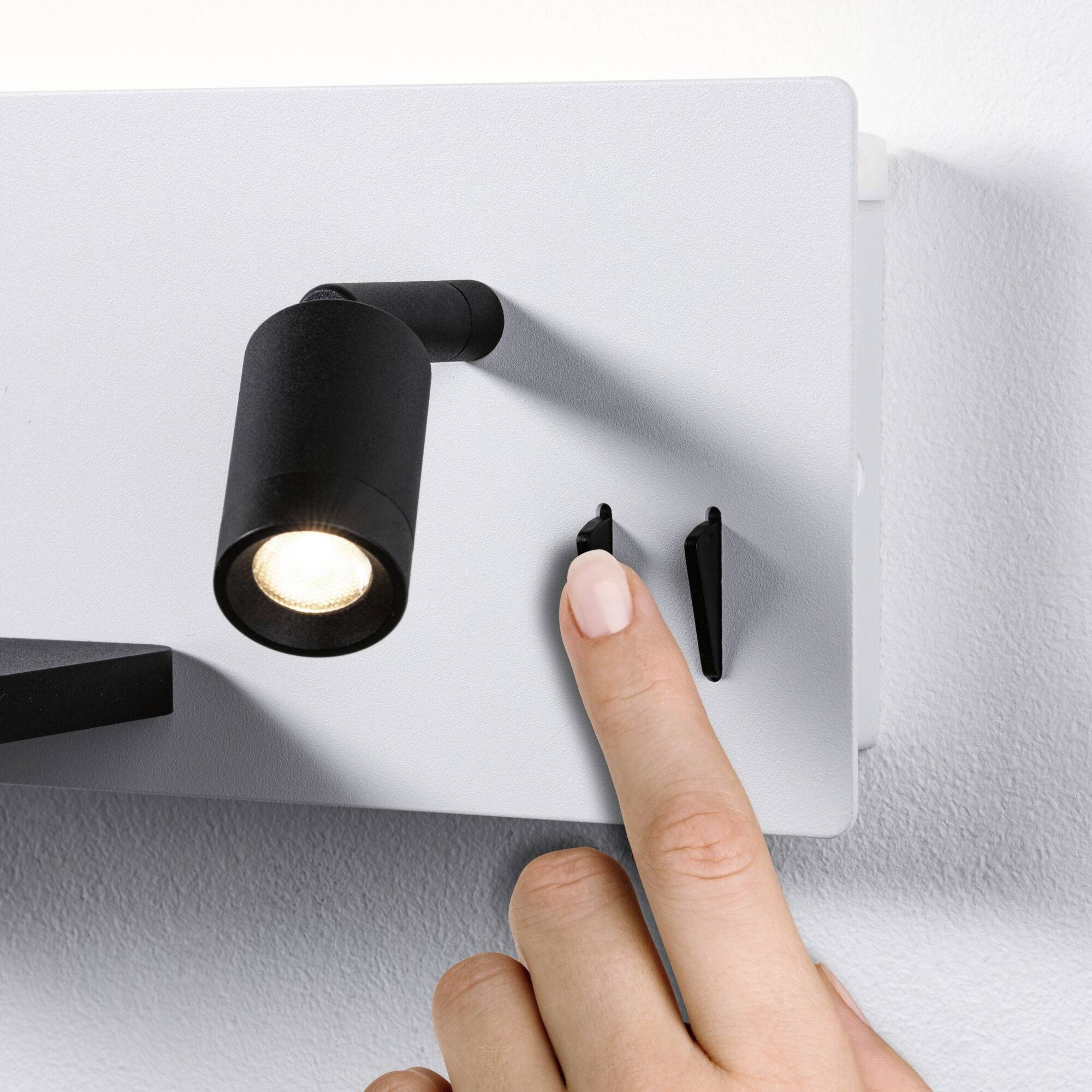Une main touche un interrupteur à côté d'un petit projecteur LED mural noir allumé qui diffuse de la lumière.