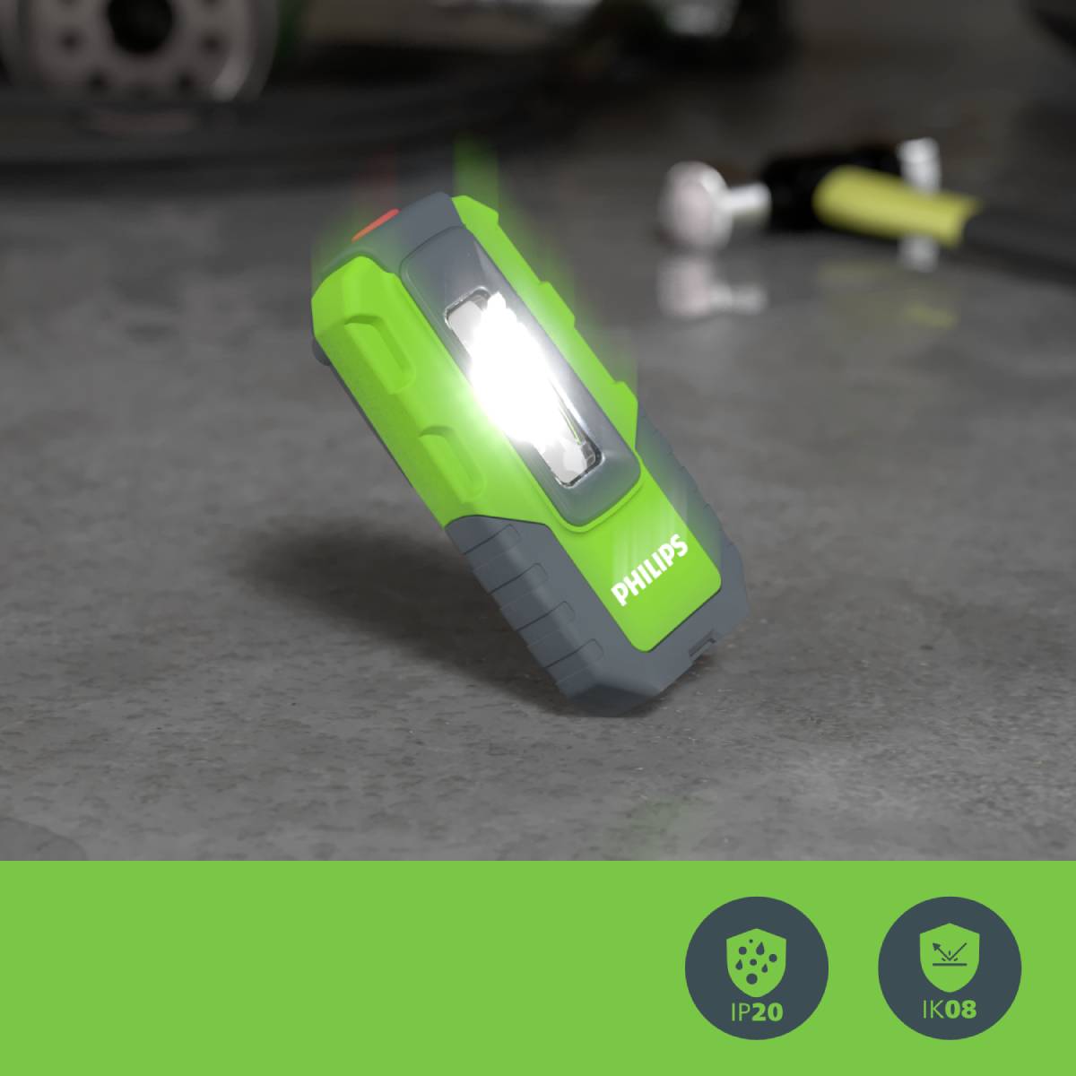 Une lampe de poche verte est posée sur un sol en béton, brillante. Des outils flous sont visibles en arrière-plan. Les symboles en bas indiquent les classes de protection IP20 et IK08.