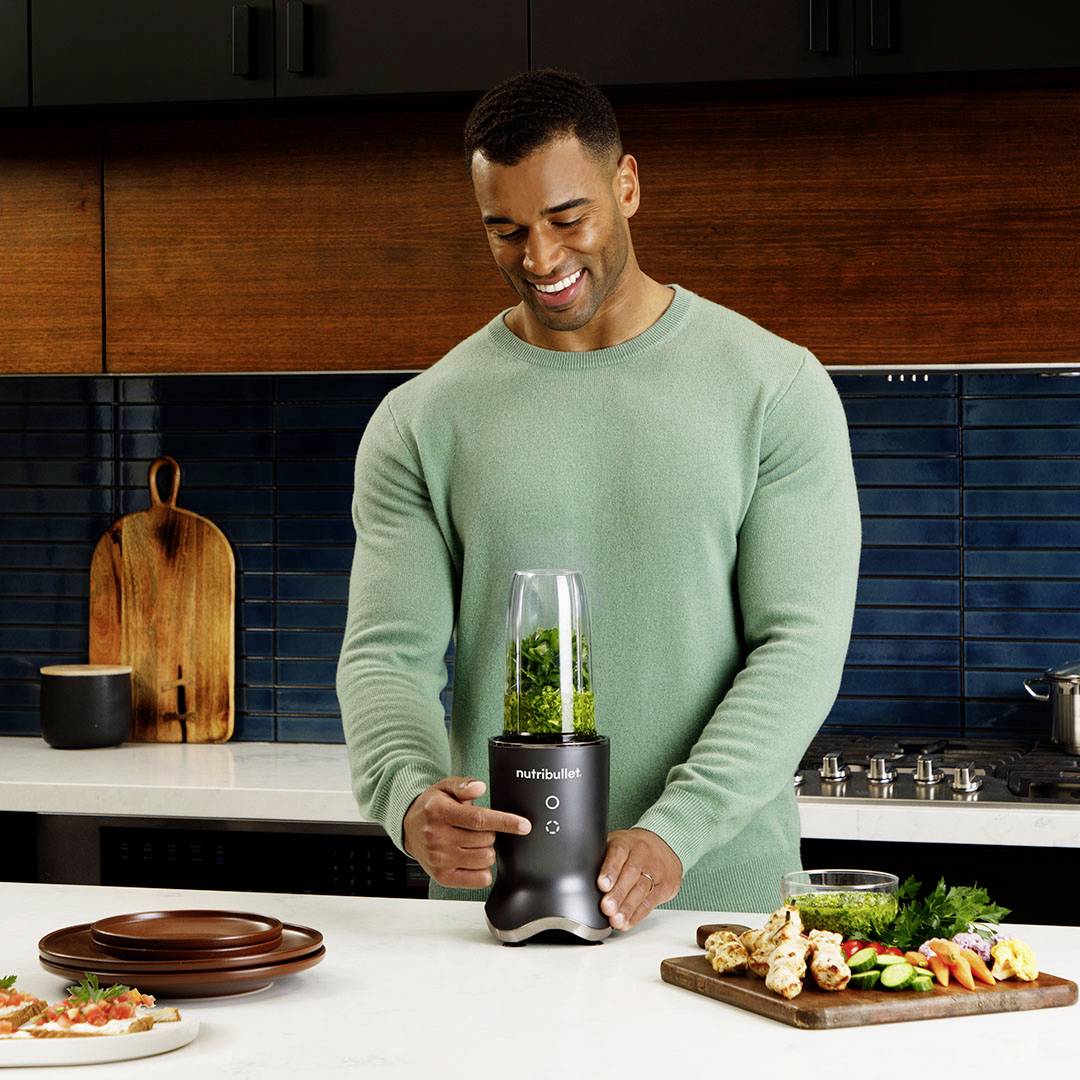 Un homme sourit tout en tenant un mixeur contenant un smoothie vert sur un plan de travail de cuisine. Des ustensiles de cuisine sont visibles en arrière-plan.