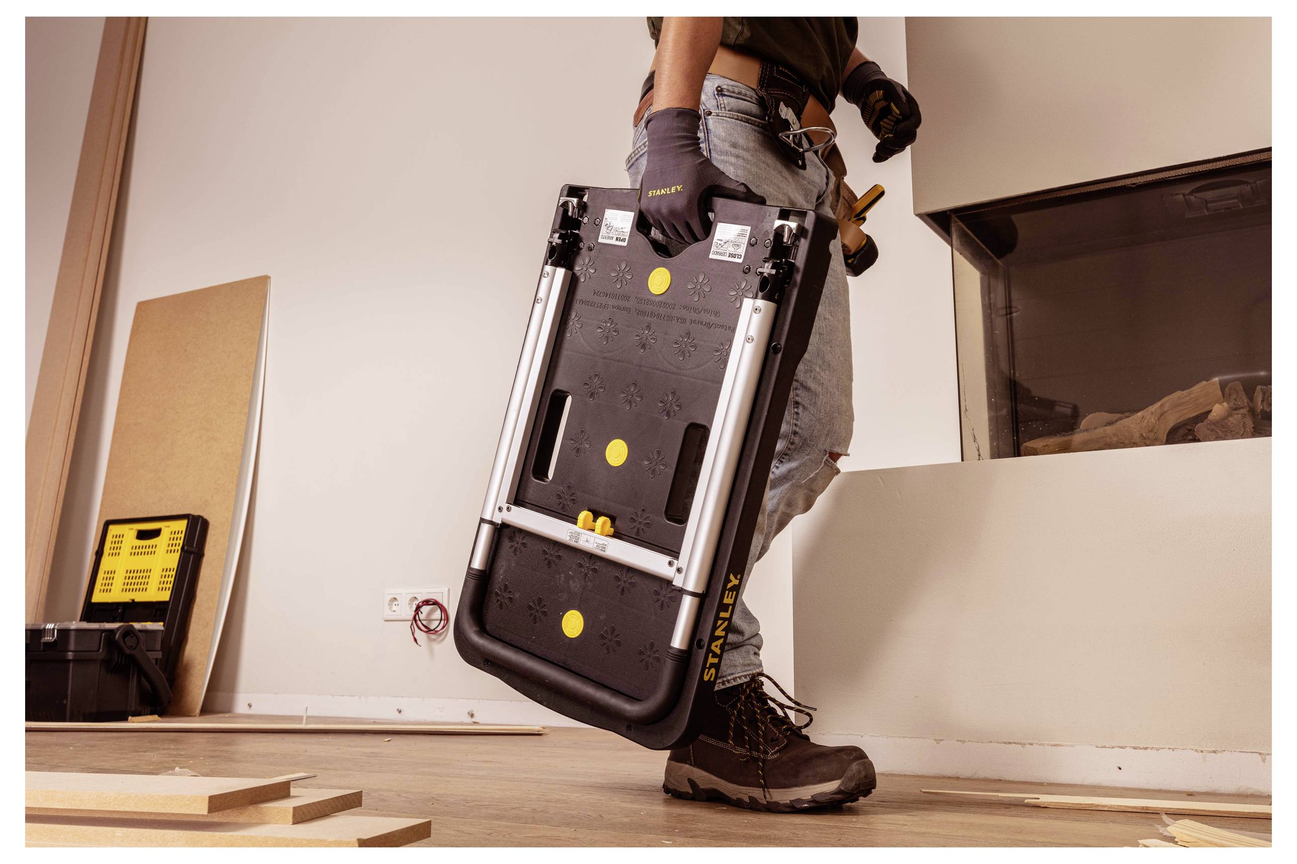 Une personne tient un établi portable plié dans un atelier, portant des gants et des bottes, ce qui suggère qu'elle se prépare pour un projet.