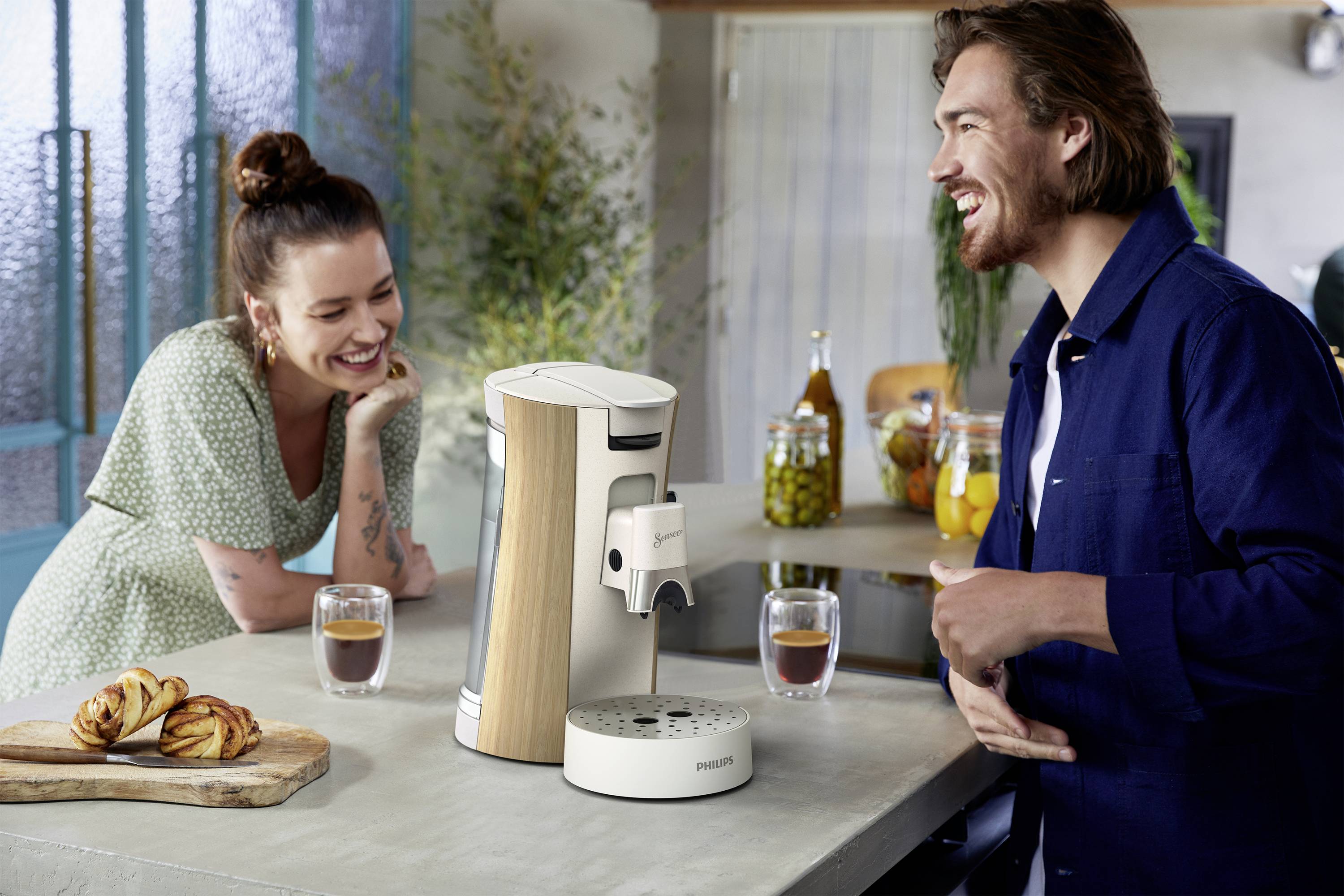 Un homme et une femme rient et discutent devant un comptoir de cuisine. Devant eux, une machine à café est posée avec deux verres de café.