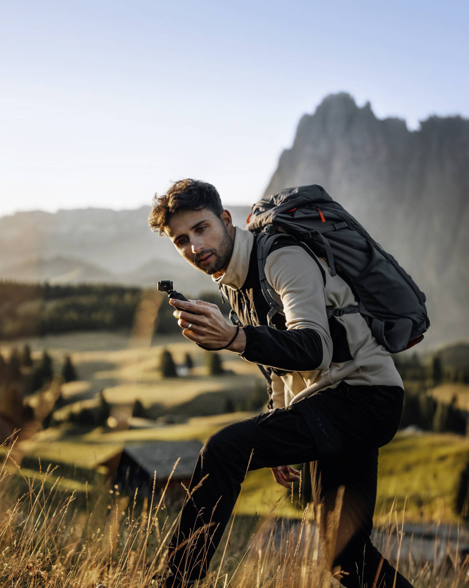 Un homme avec un sac à dos tient un appareil photo dans un paysage montagneux. Le soleil du matin illumine les prairies et les montagnes en arrière-plan.