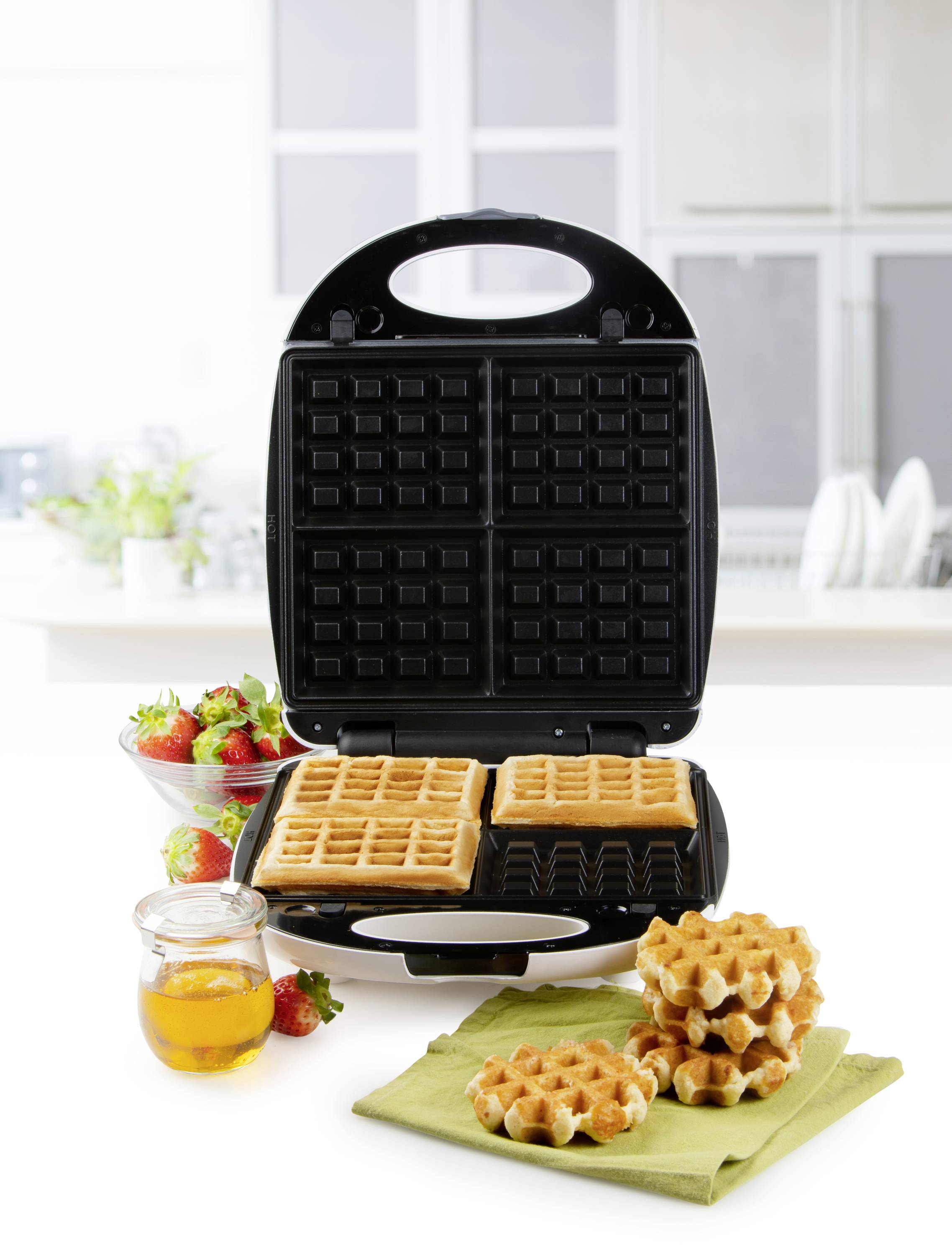 Un distributeur de gaufres ouvert rempli de gaufres dorées, entouré de fraises fraîches, de miel et d'une pile de gaufres sur un torchon.