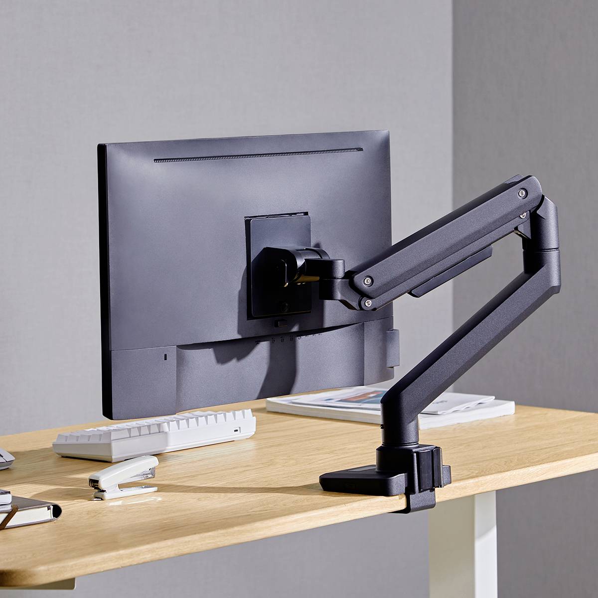 Un écran d'ordinateur équipé d'un bras de support flexible et ergonomique est installé sur un bureau qui contient également un clavier et des livres.