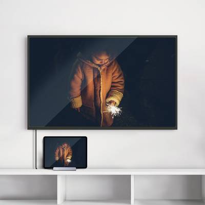 Un enfant portant un manteau d'hiver se tient dans l'obscurité et tient un cierge magique lumineux. L'image est visible sur un téléviseur et une tablette.