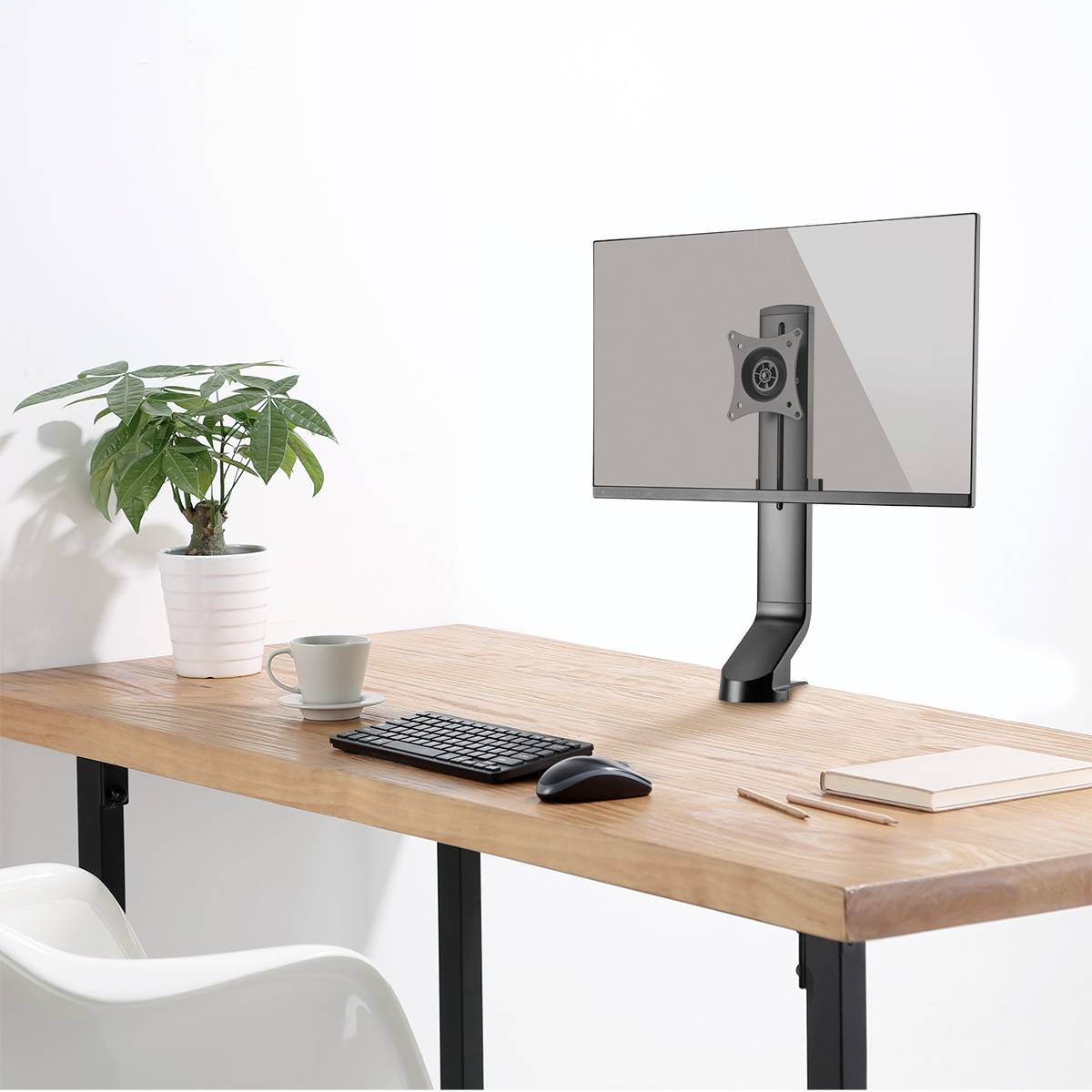 Sur un bureau se trouve un support de moniteur sans écran. À côté, il y a un clavier, une souris, un livre et une plante dans un pot.