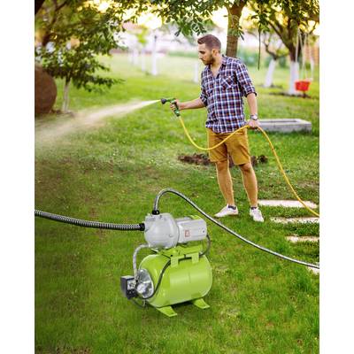 Homme portant une chemise à carreaux et un short, arrosant la pelouse avec un tuyau d'arrosage. Au premier plan, une pompe à eau verte. En arrière-plan : des arbres verts.