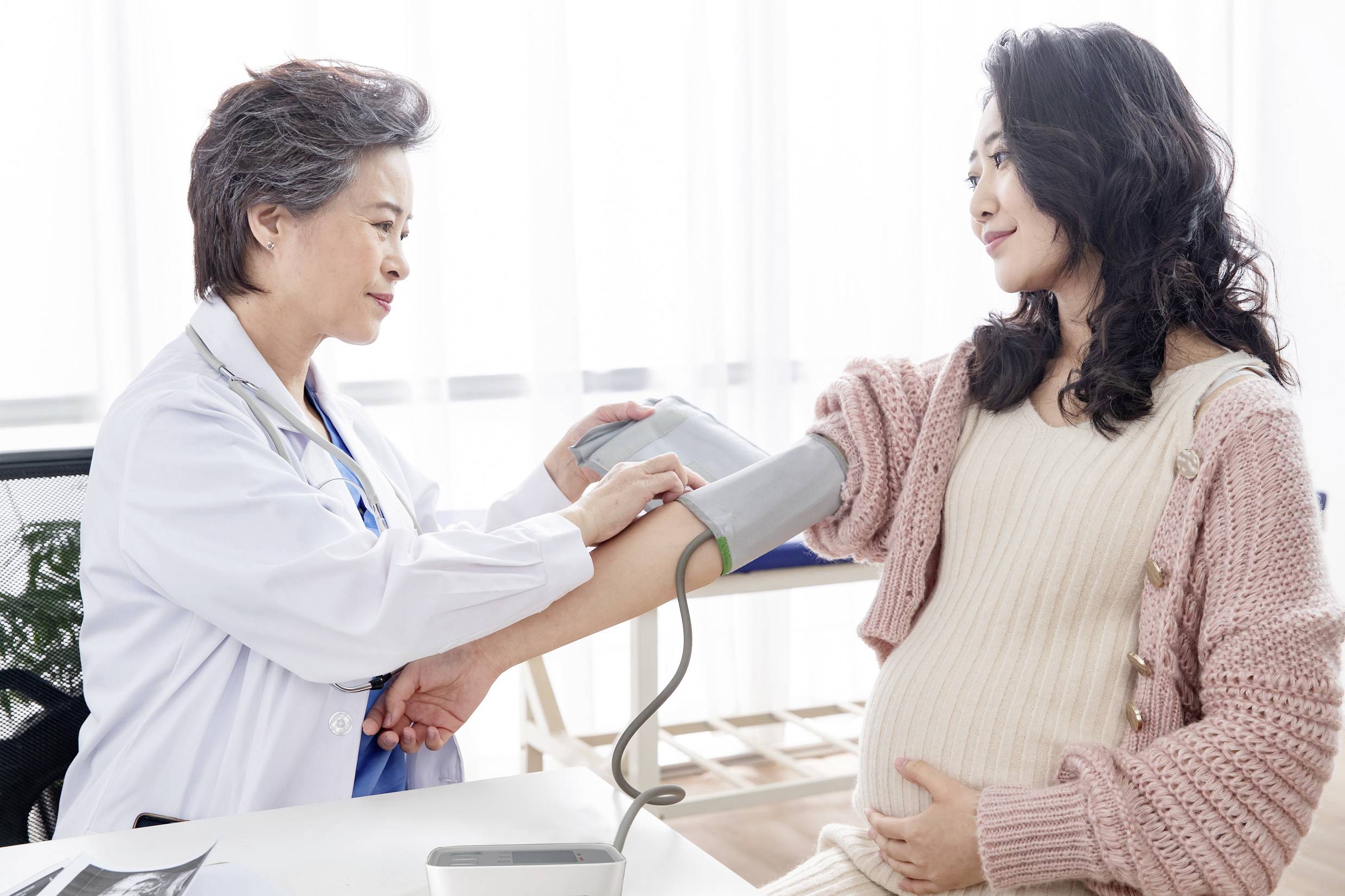 Une médecin mesure la tension artérielle d'une femme enceinte. La femme est assise sur une chaise et tient son ventre.