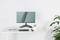 Un moniteur d'ordinateur moderne sur un bureau blanc. À côté se trouvent un clavier, un livre ouvert, un porte-stylos et une plante.