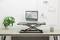 Un convertisseur de bureau réglable en hauteur sur lequel est posé un ordinateur portable, entouré d'une plante de bureau, de notes accrochées à une grille et d'accessoires de bureau.
