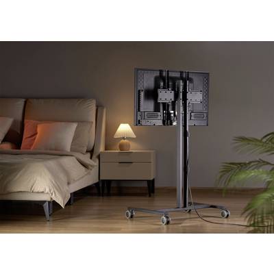 Une chambre à coucher avec un lit à gauche, une table de chevet avec une lampe derrière et un support de télévision mobile à droite sur un sol en bois.
