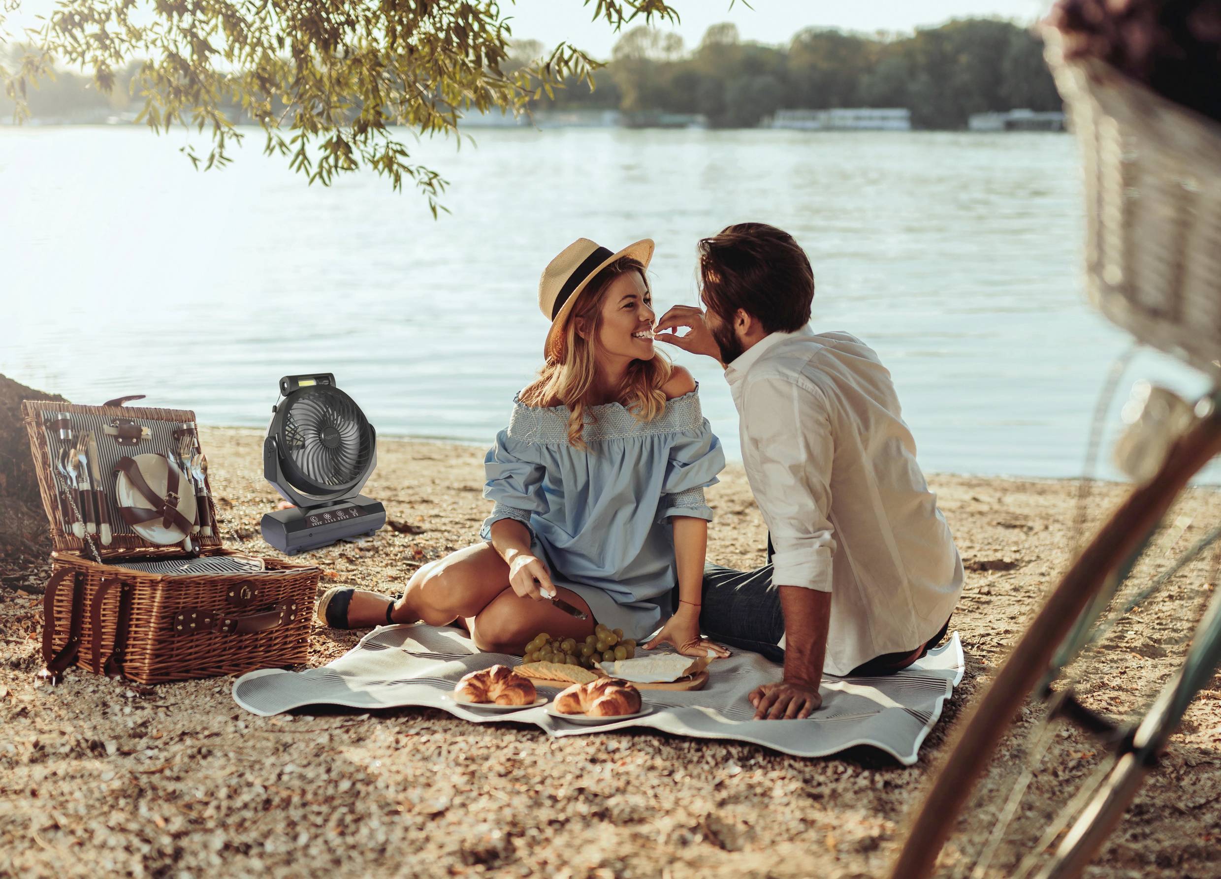 Un couple est assis sur une couverture au bord de la rivière pour un pique-nique. Ils rient et mangent des sandwichs. En arrière-plan, on aperçoit une rivière.