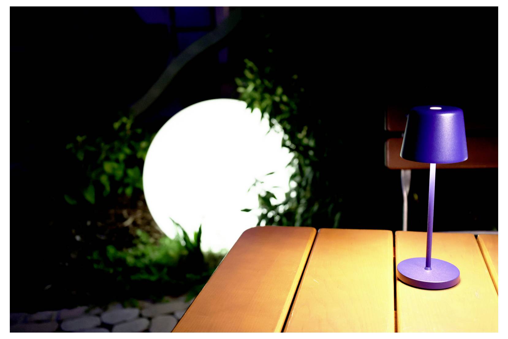 Lampe de table bleue posée sur une table en bois en extérieur, la nuit, éclairée par un grand luminaire rond de jardin en arrière-plan.