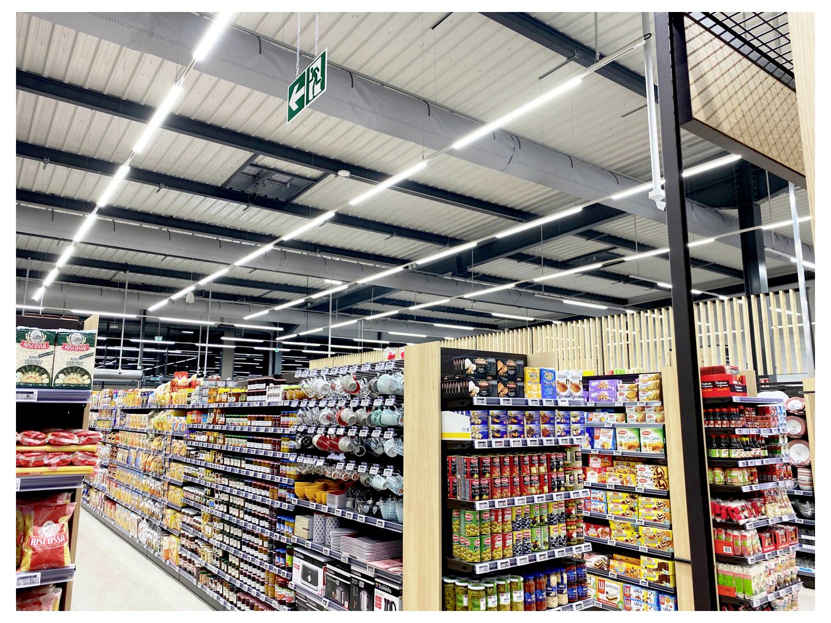 Une allée de supermarché très éclairée, bordée d'étagères de produits alimentaires emballés, de conserves et de bouteilles, avec vue sur une enseigne de sortie verte au-dessus.