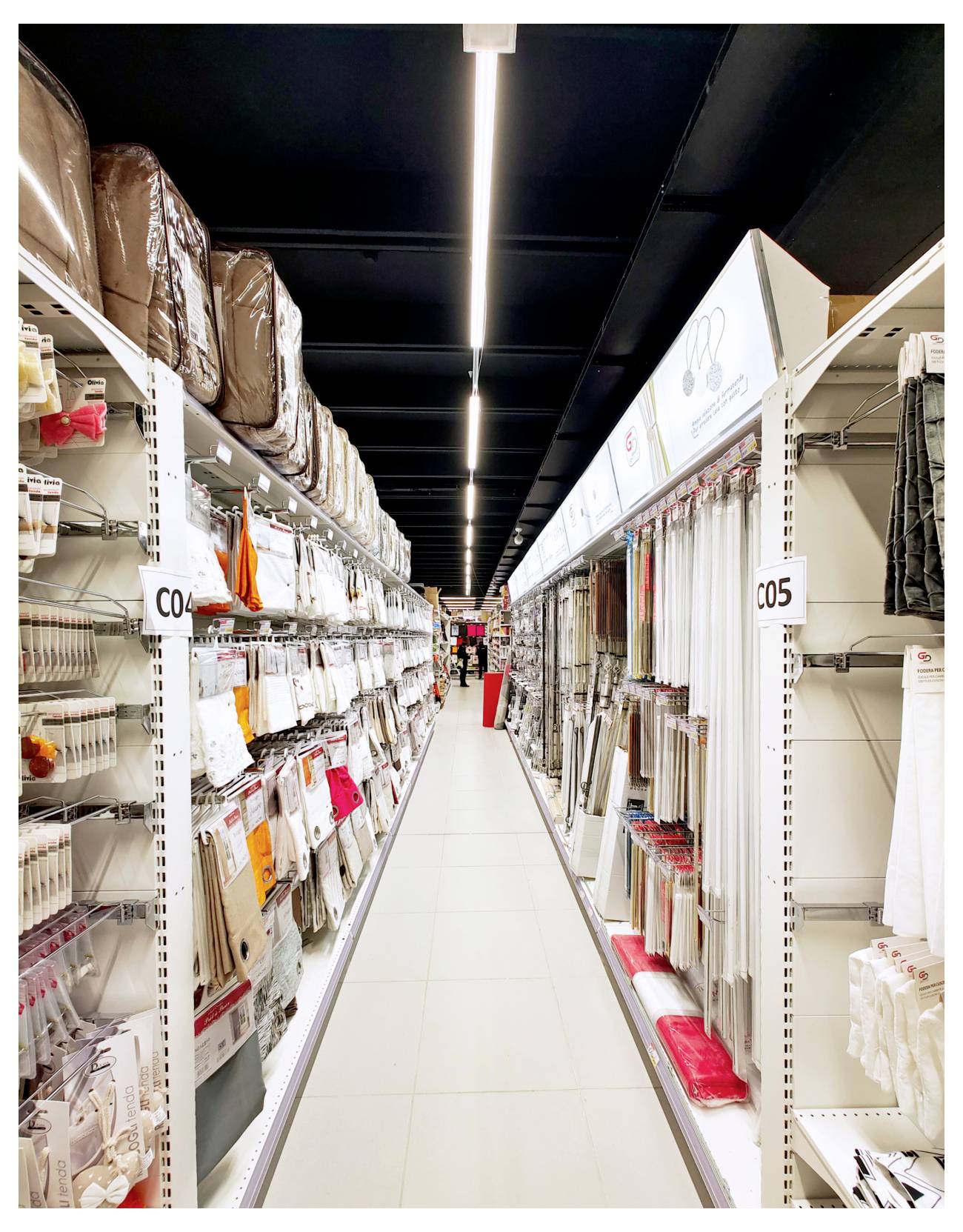 Une allée de magasin bien éclairée, bordée de rayonnages garnis de textiles pliés, de sacs et d'articles pour la maison, signalée par les étiquettes de rayon C04 et C05.