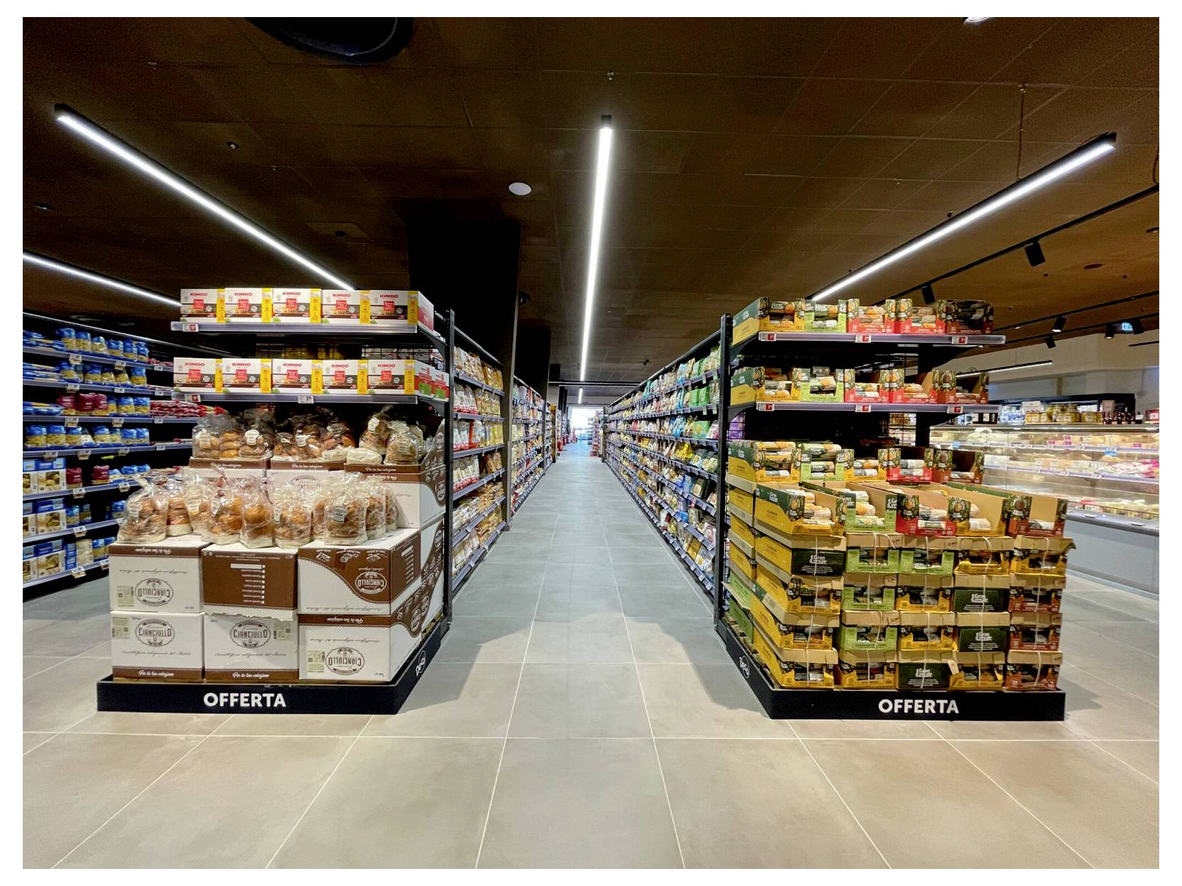 Allée de supermarché avec deux présentoirs promotionnels en bout de rayon étiquetés « OFFERTA », garnis de produits emballés et de pain sous des plafonniers lumineux.