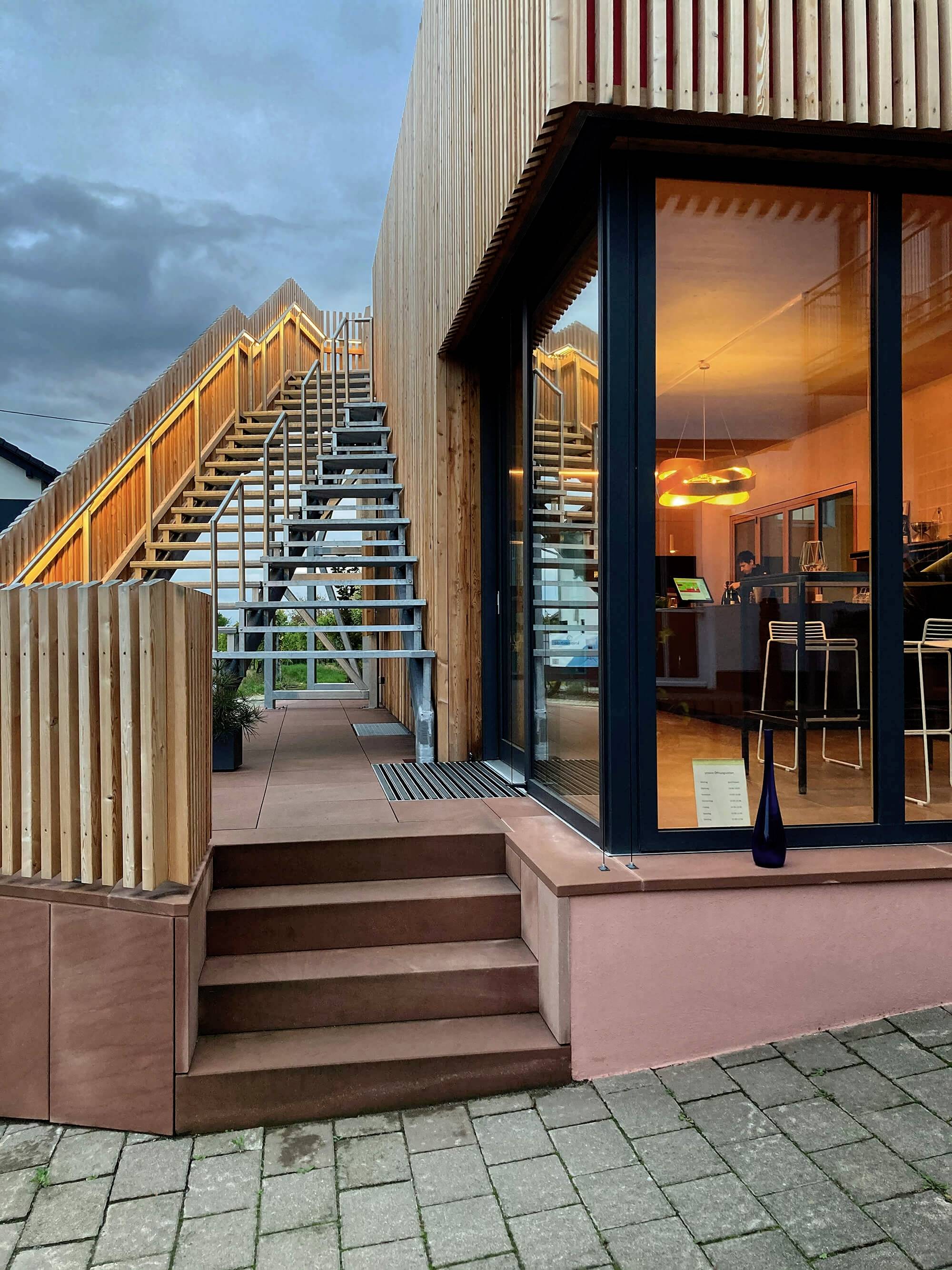 Bâtiment moderne avec un revêtement en bois, de grandes fenêtres vitrées et un escalier extérieur. L'intérieur présente un aménagement minimaliste.